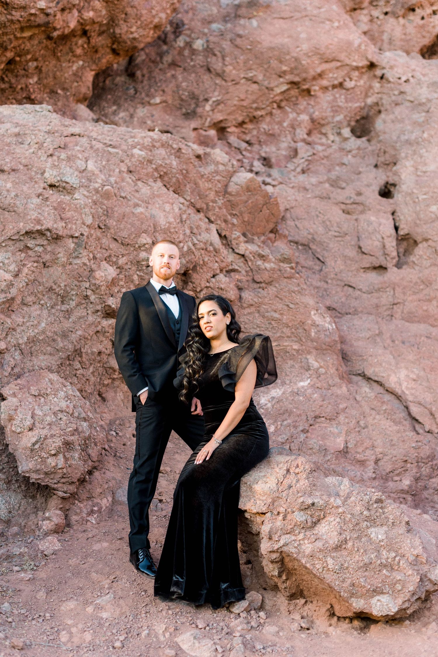 Formal portrait of couple in black attire against red rock wall.