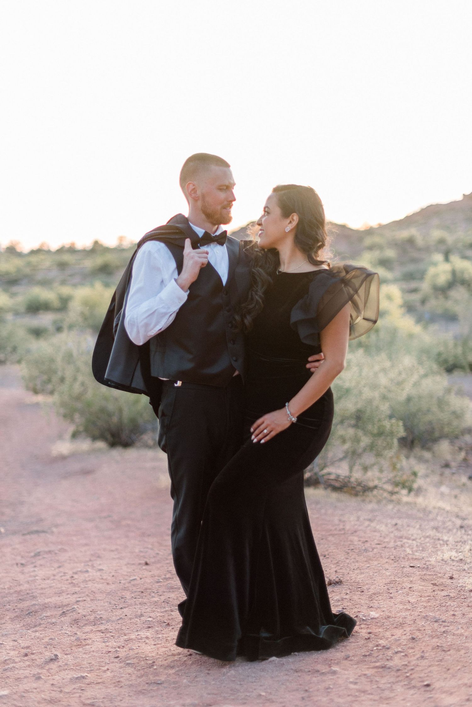 Couple in formal wear shares intimate moment on desert trail at sunset.
