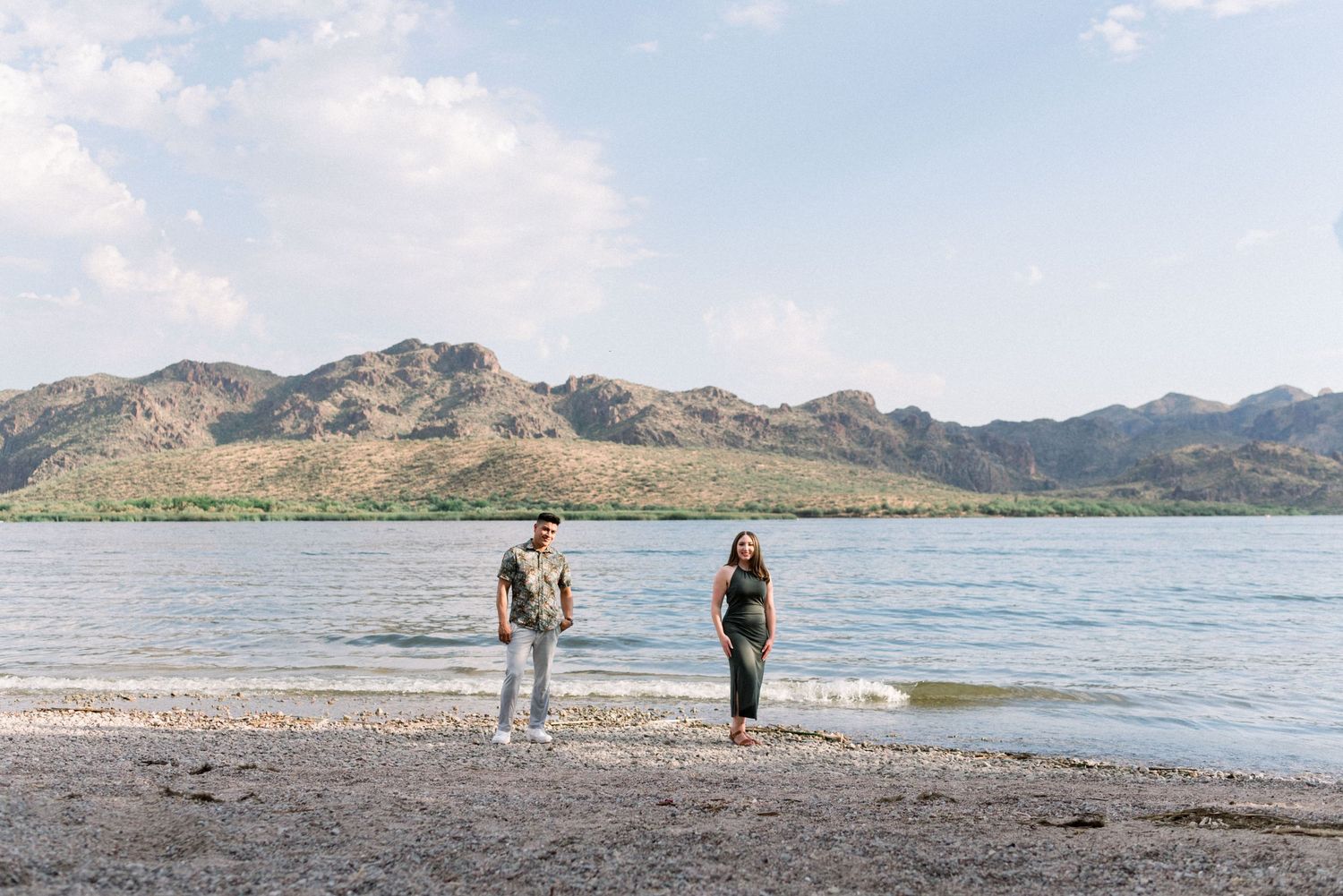 A couple walks along a scenic beach with mountains and blue sky in the background.