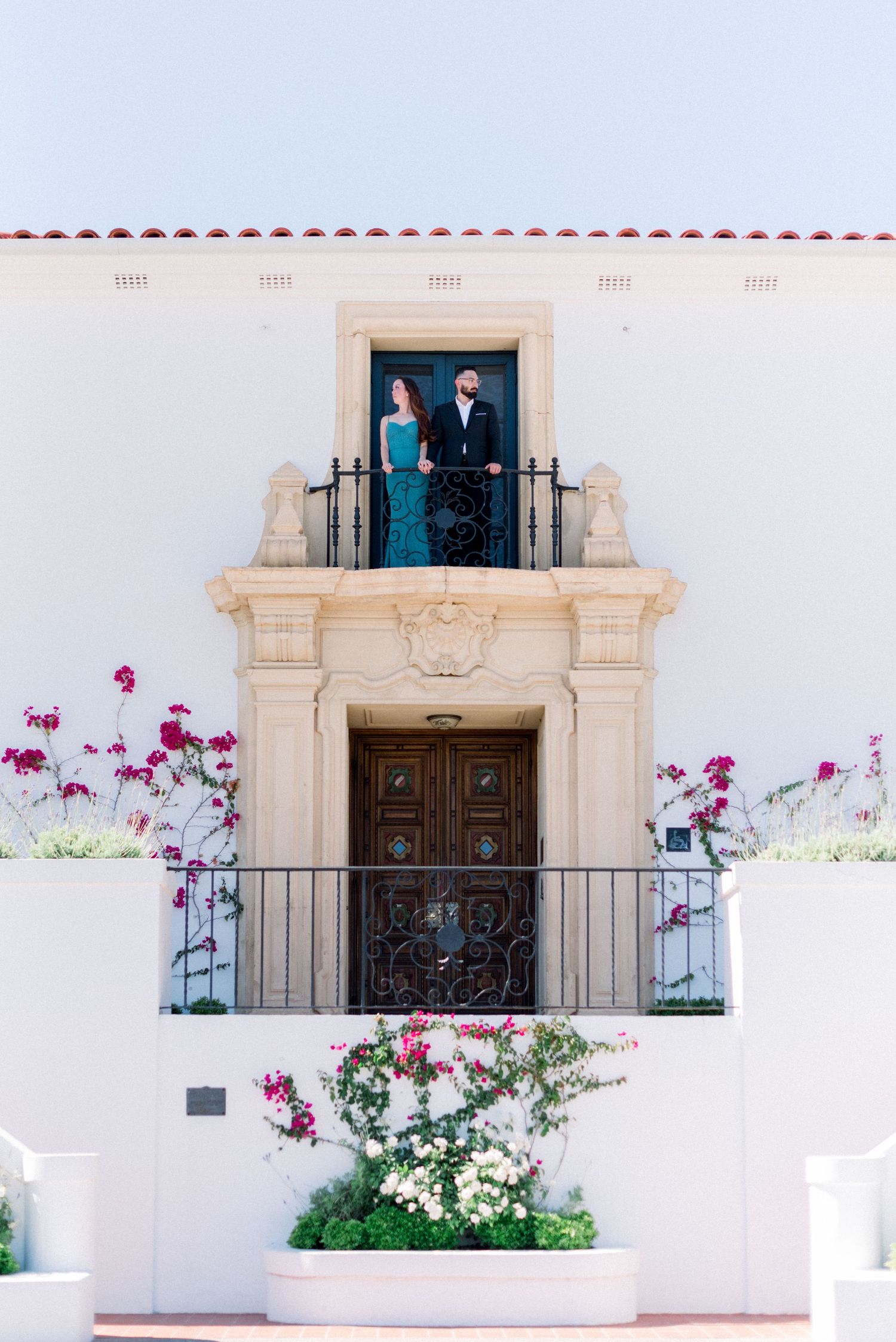 Couple stands in ornate doorway of Mediterranean-style building with pink flowers.