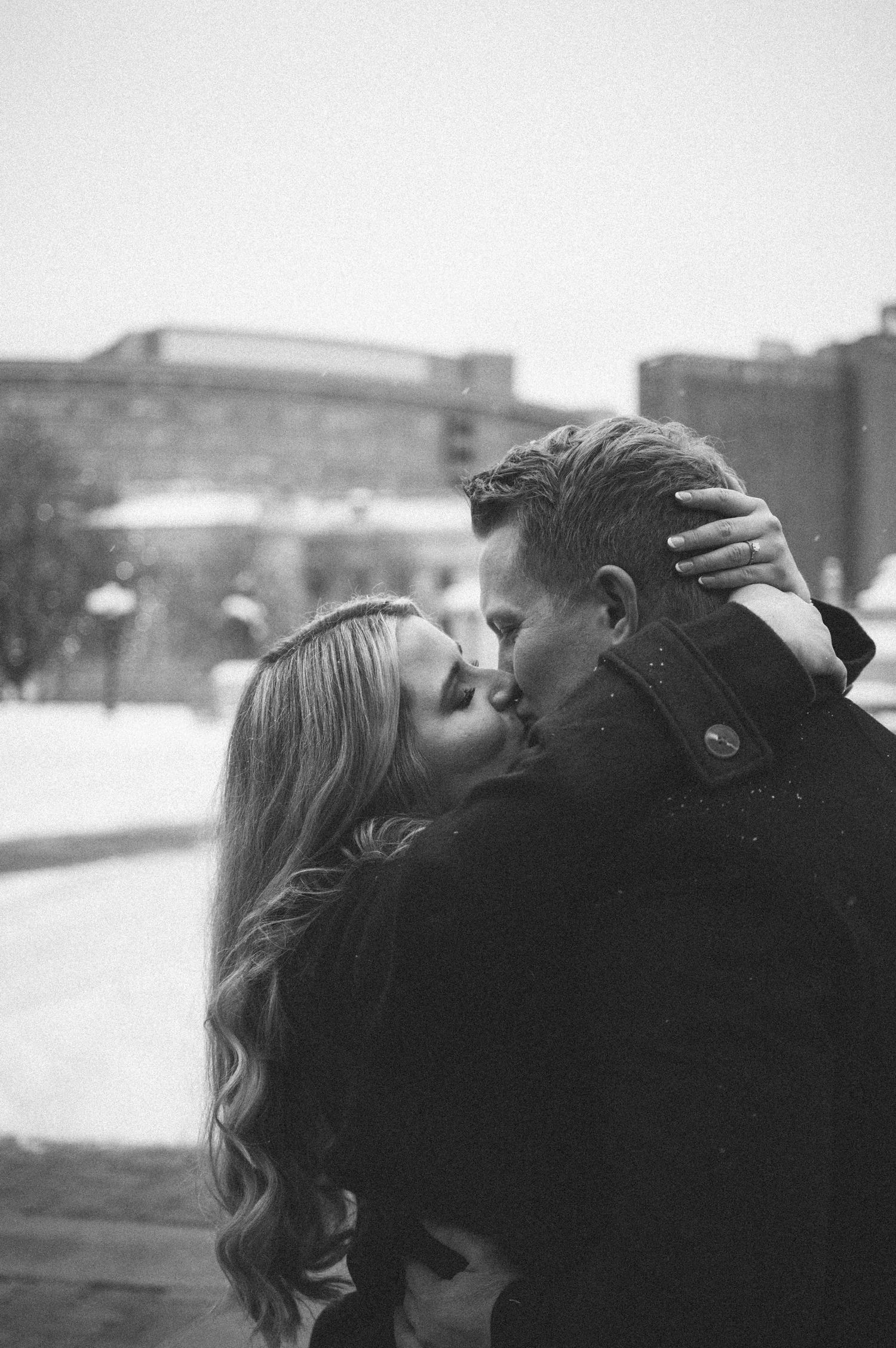 A romantic winter kiss between a couple against a snowy urban backdrop.