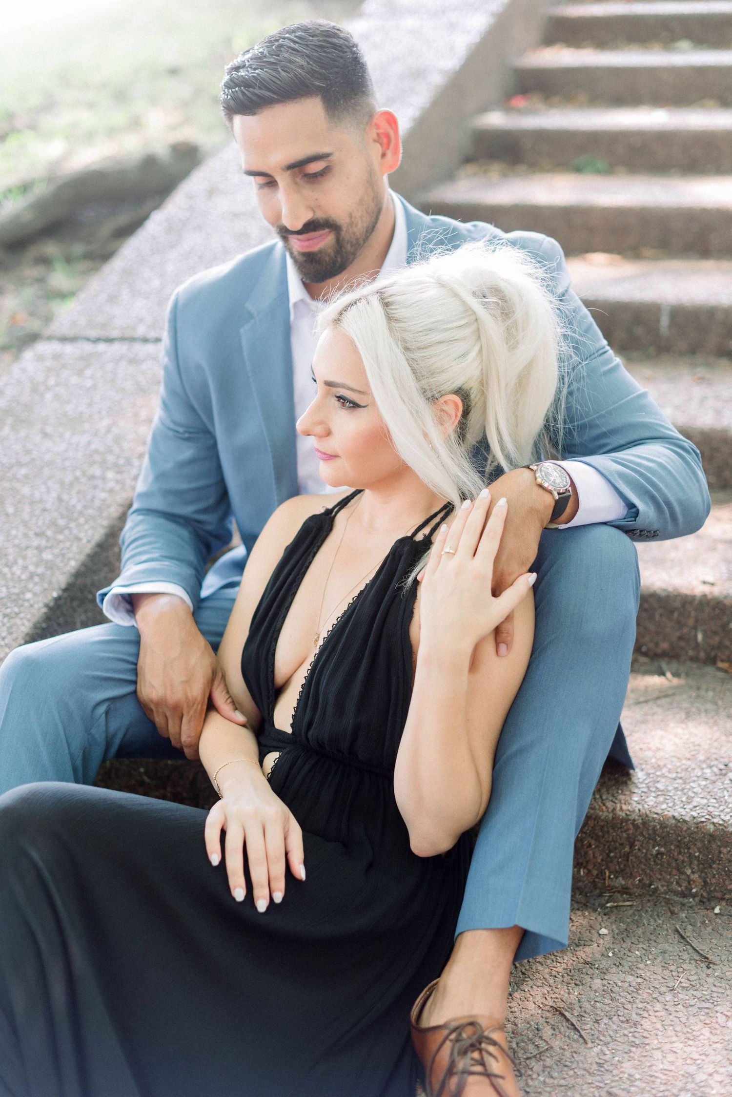 Romantic couple in blue suit and black dress embracing while seated on concrete steps.