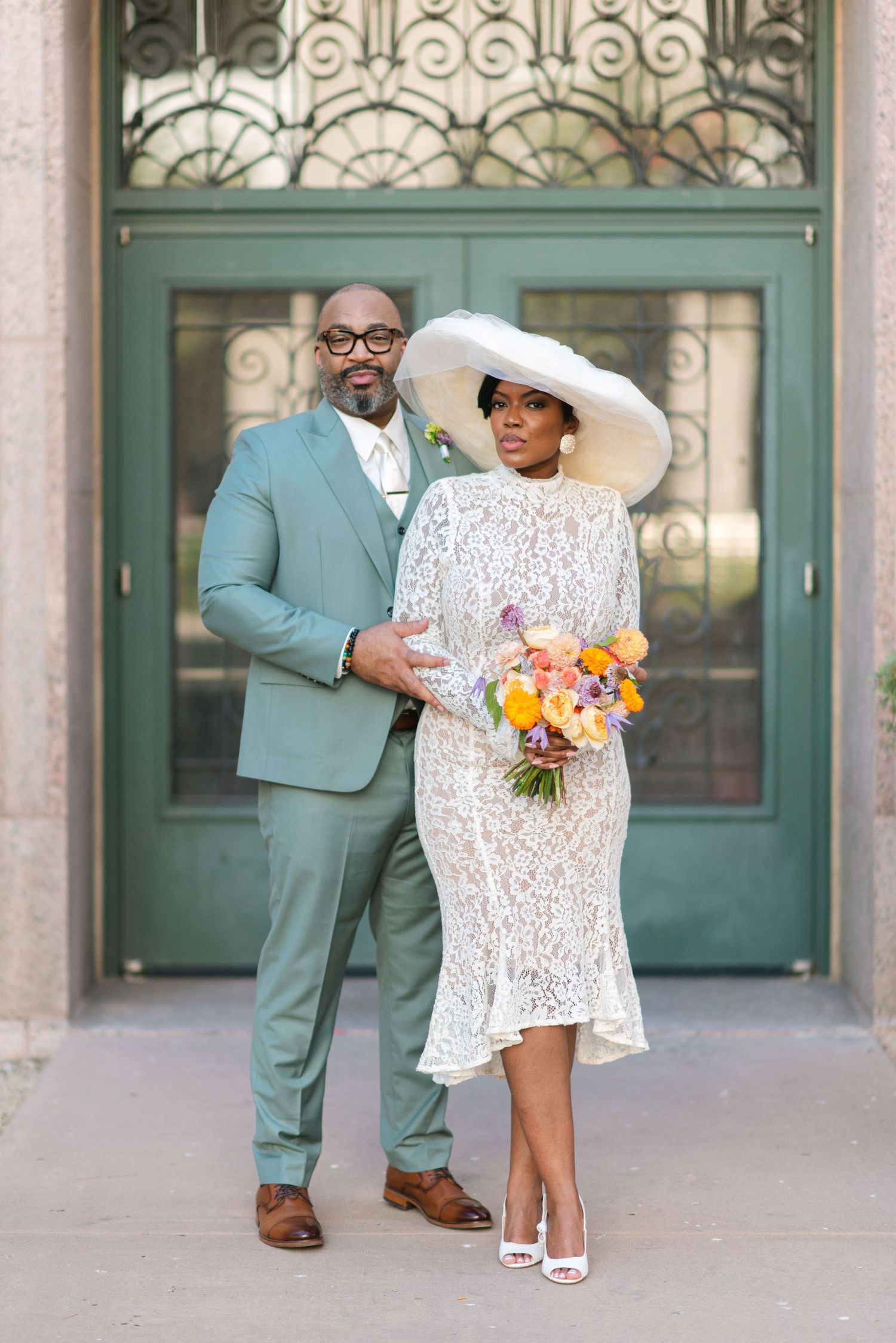 Elegant couple in mint green suit and white lace dress with wide brim hat pose in front of ornate doorway.