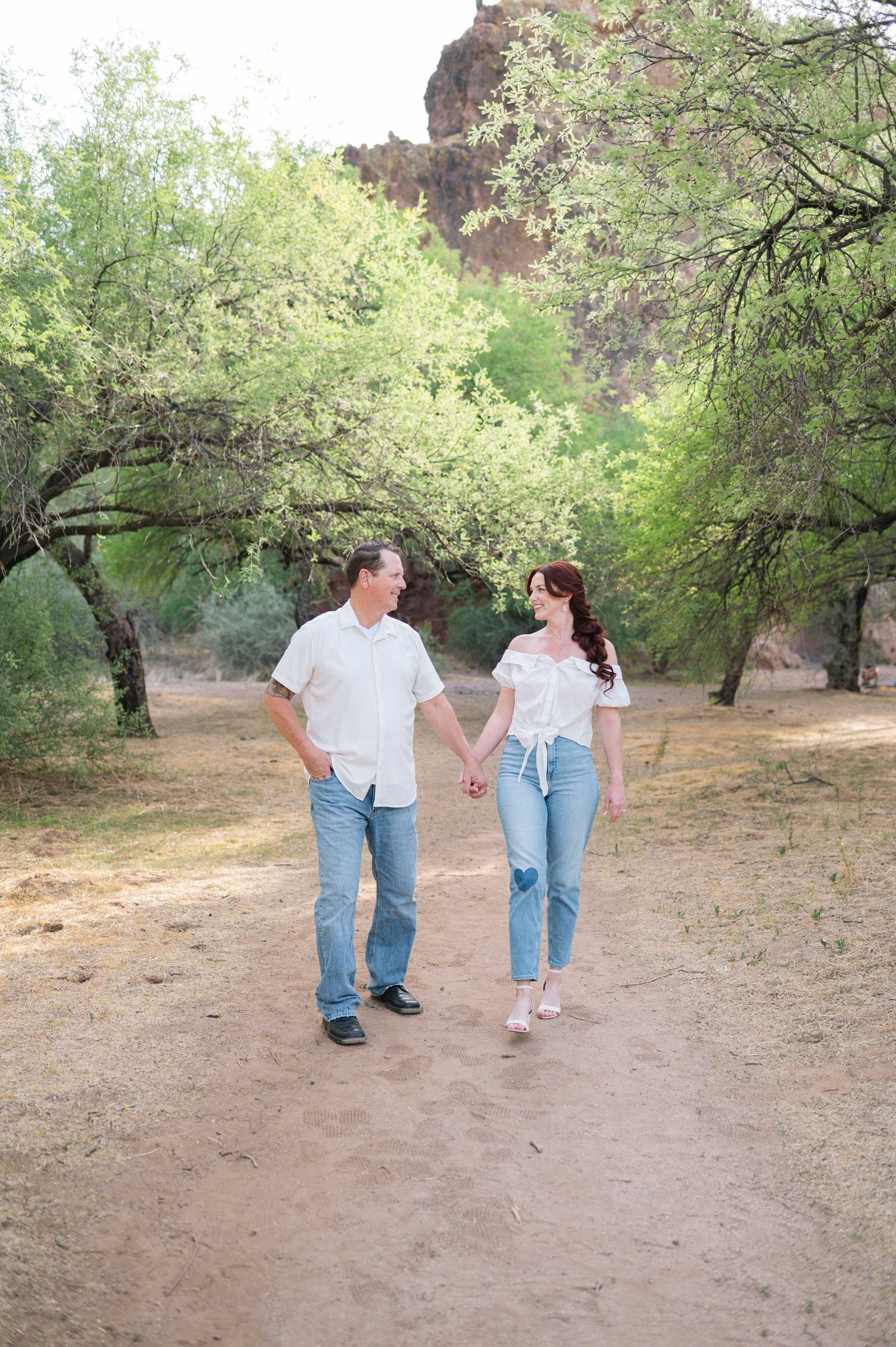 A couple walking hand in hand along a dirt path through a grove of trees in a natural outdoor setting.