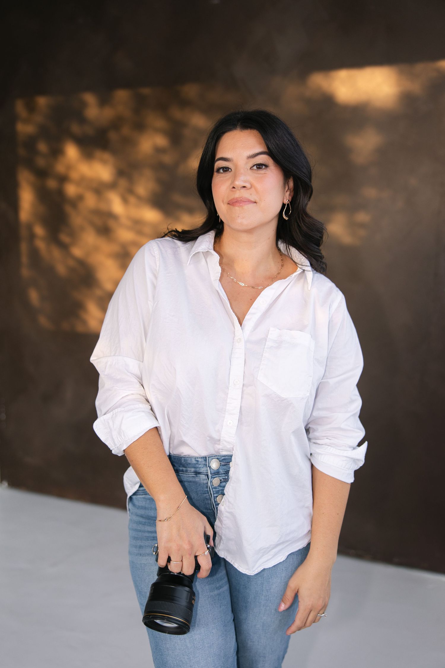 Professional photographer in white button-down shirt and jeans poses with camera equipment against dark backdrop.