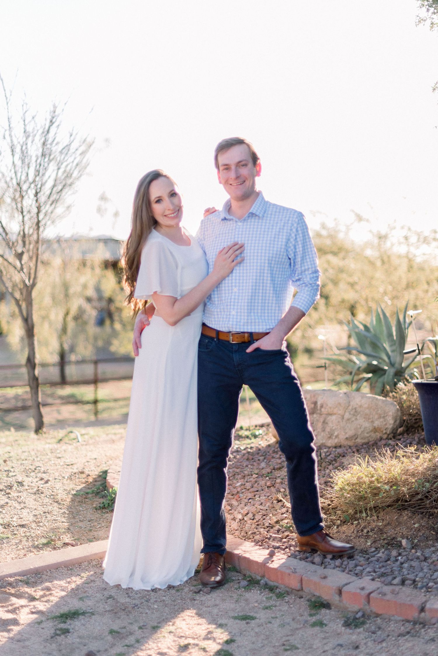 Engaged couple poses in desert garden wearing white dress and blue checkered shirt.