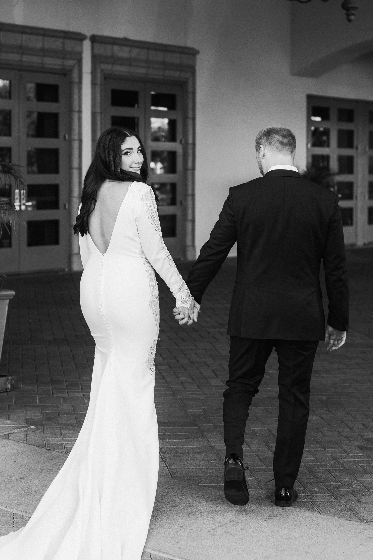 Back view of couple walking hand in hand, bride in backless gown and groom in classic tuxedo.