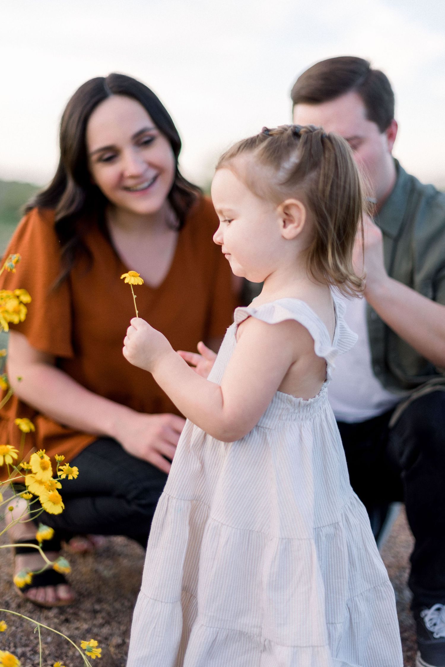 Child in white dress examines yellow wildflower in desert setting.