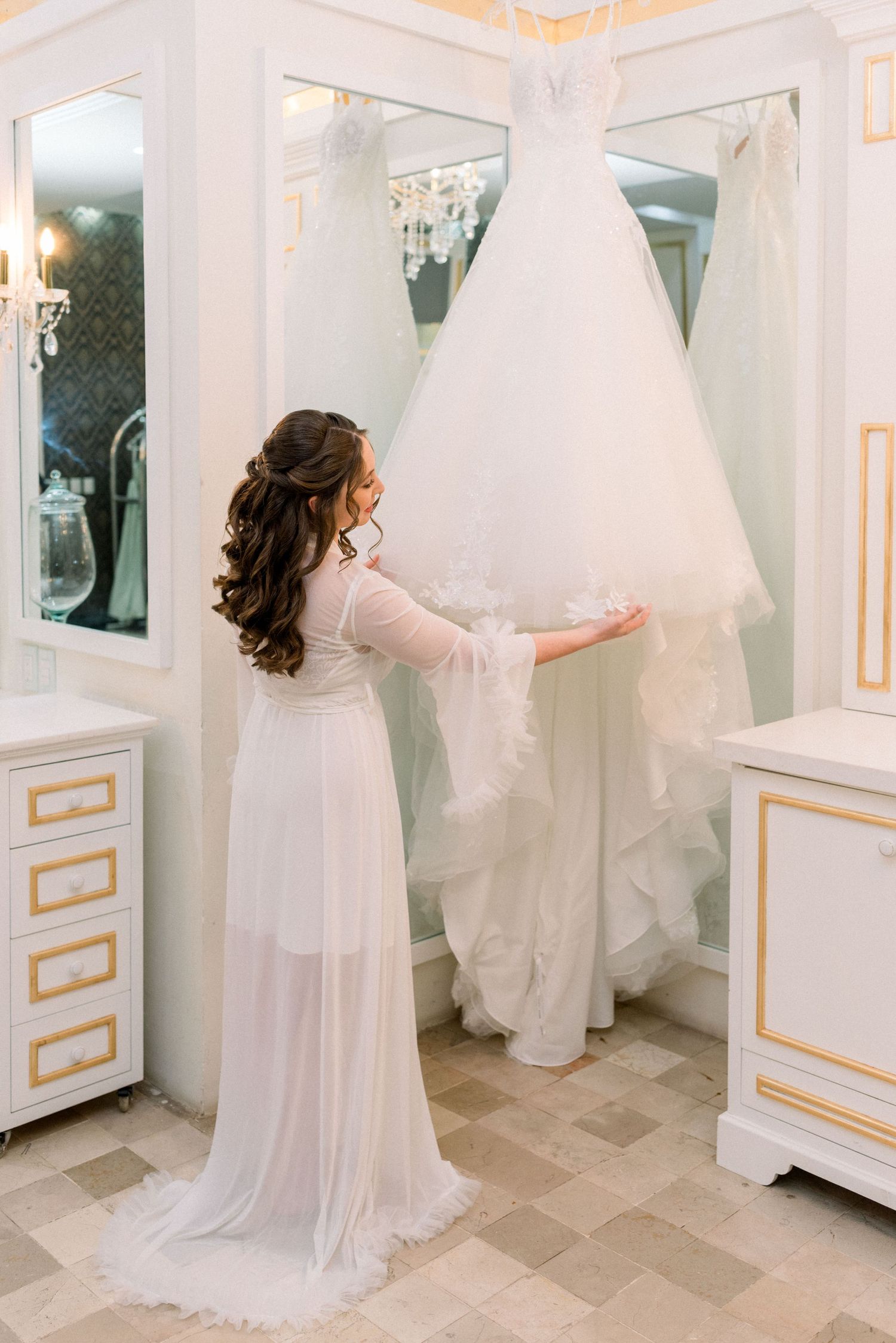 Wedding dress preparation scene in an elegant white dressing room with mirrors and gold accents.