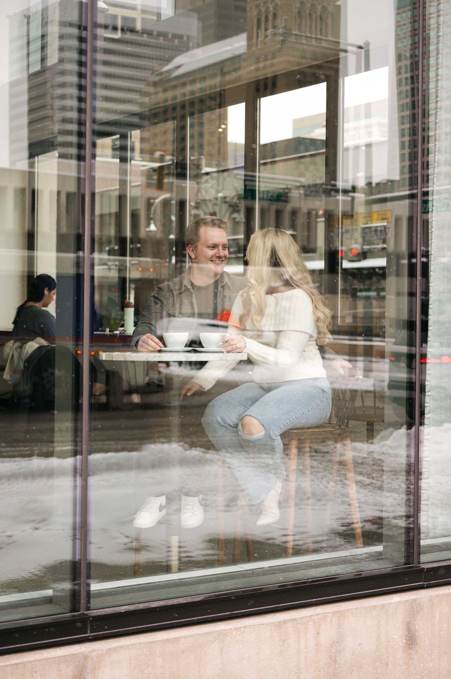 A couple enjoys coffee inside a cafe viewed through large glass windows with city reflections.