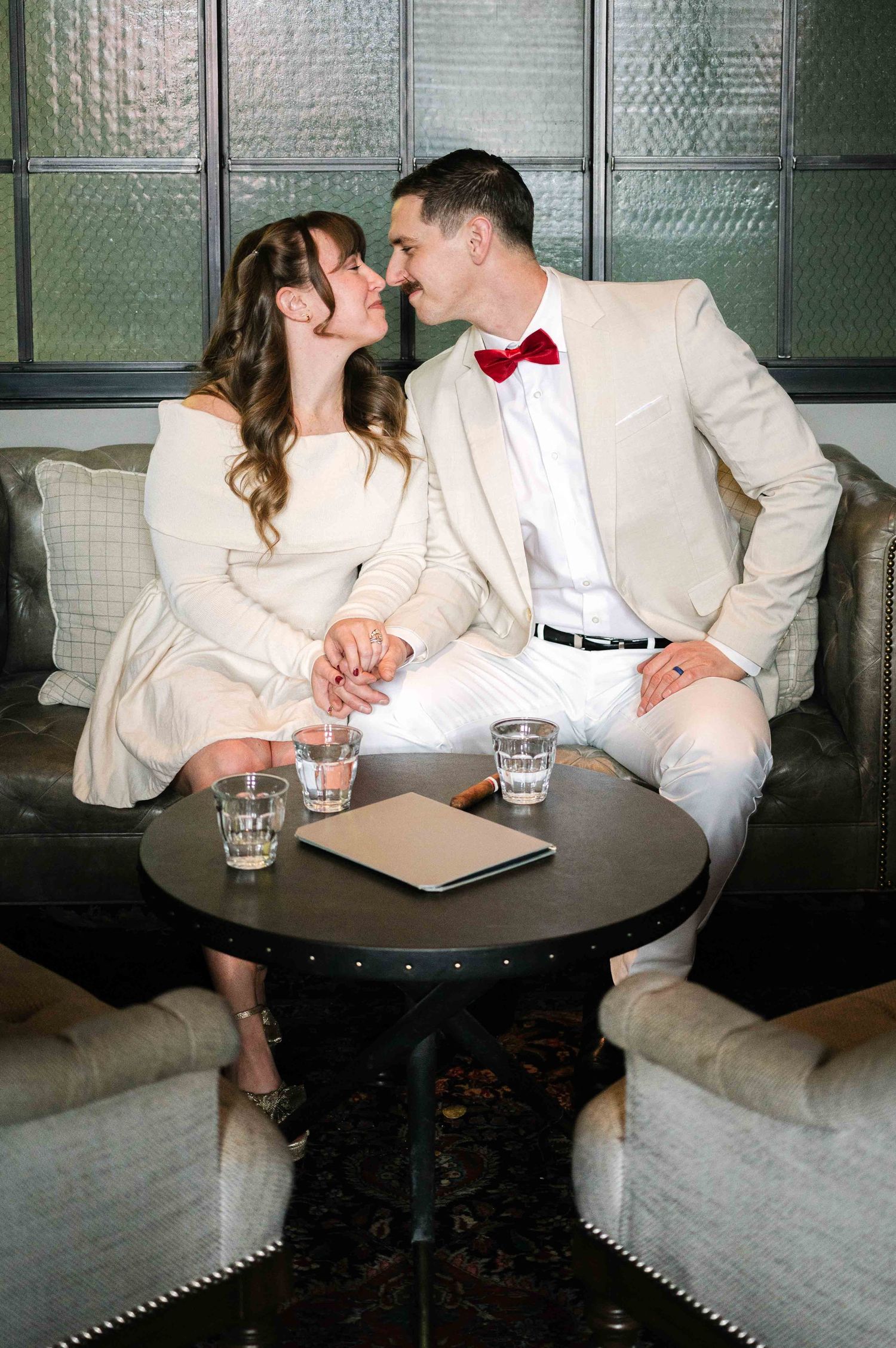 A romantic couple in cream outfits sharing an intimate moment at a lounge table with glasses.