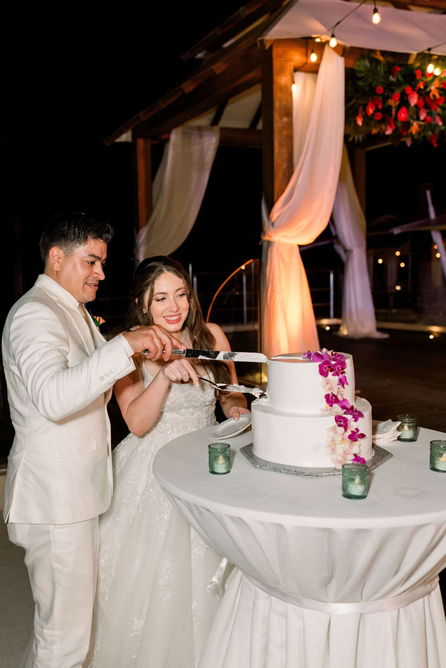Newlyweds cut their white wedding cake together under a decorated gazebo with flowing fabric and lights.
