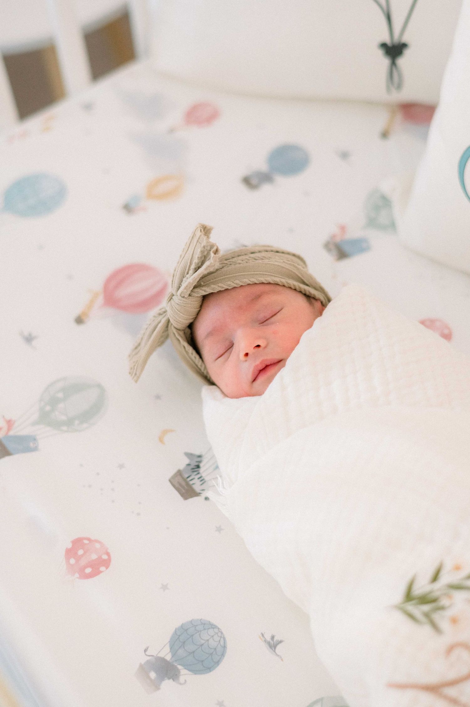 Sleeping newborn with beige bow headband on balloon-patterned bedding.