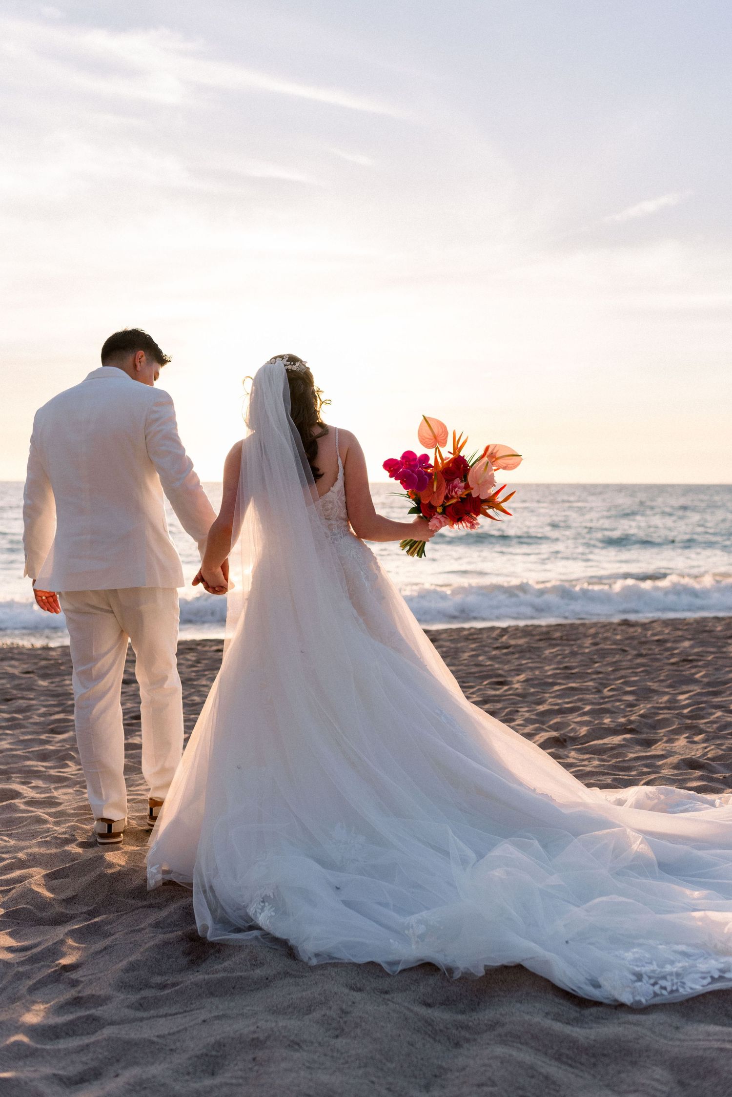 Newlyweds holding hands on beach at sunset with bride holding vibrant tropical bouquet and flowing white dress train.
