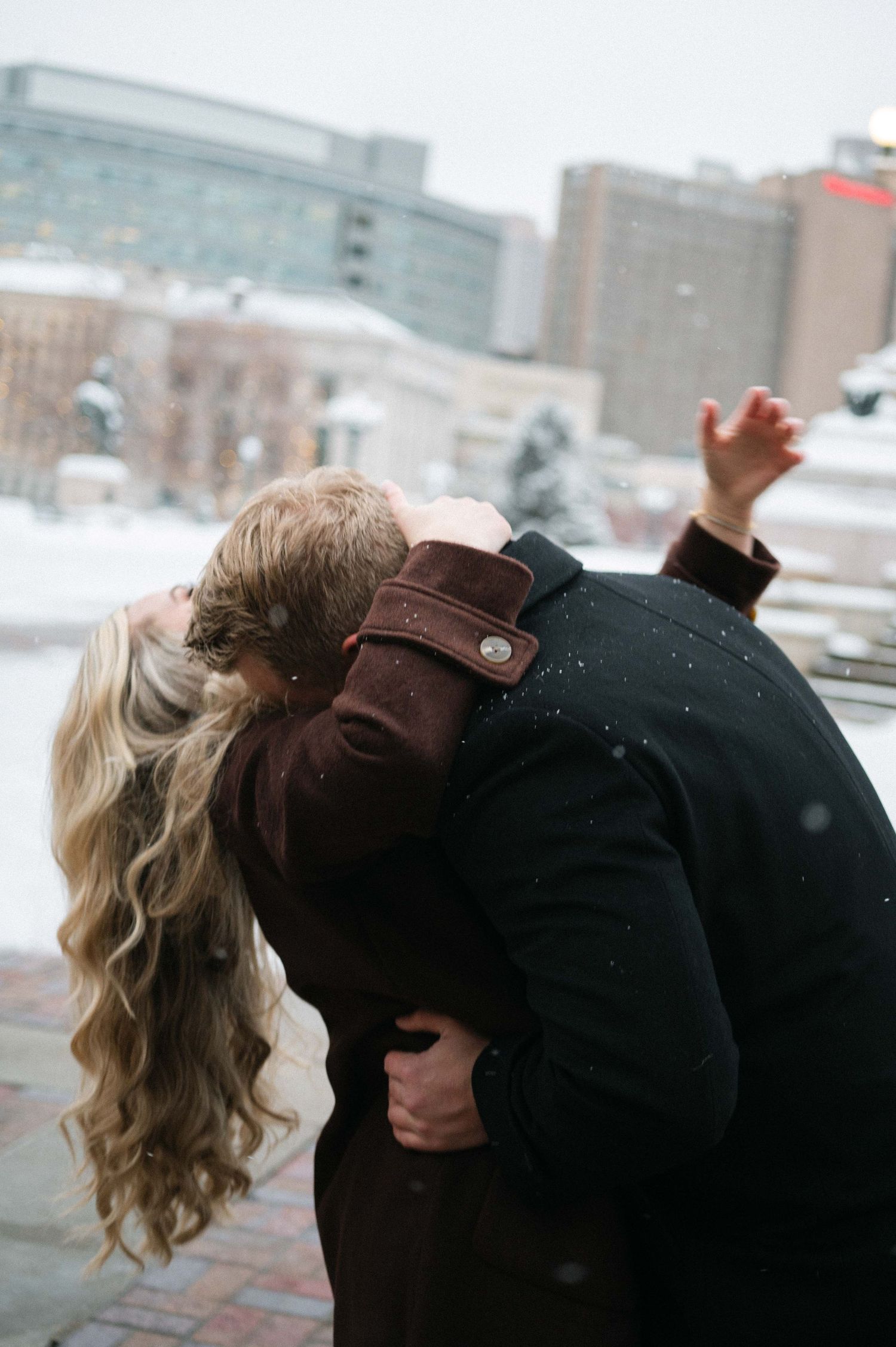 A romantic winter moment captured with city buildings in the background during snowfall.