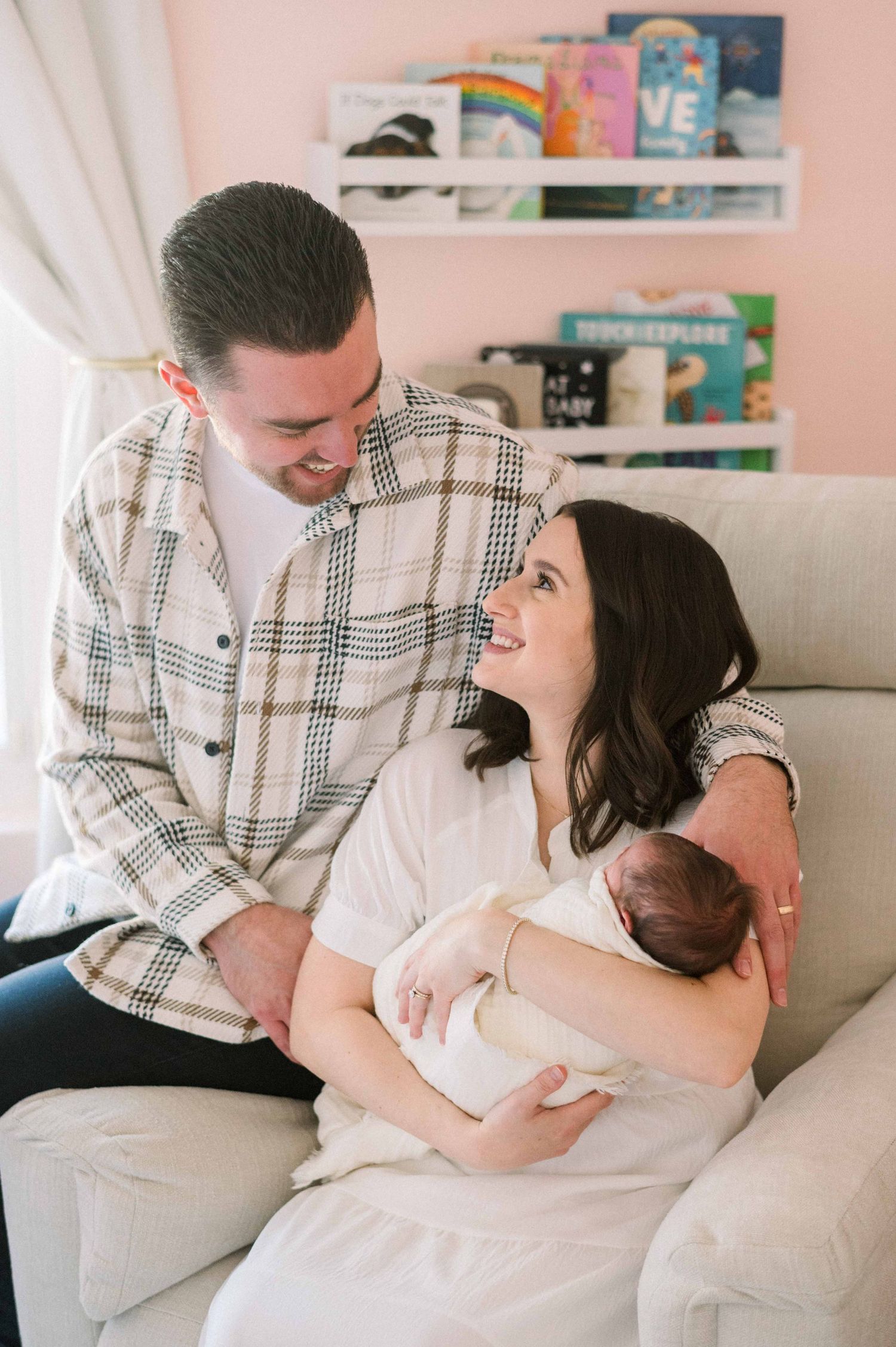 A couple shares a joyful moment on a couch while holding their newborn in a nursery.