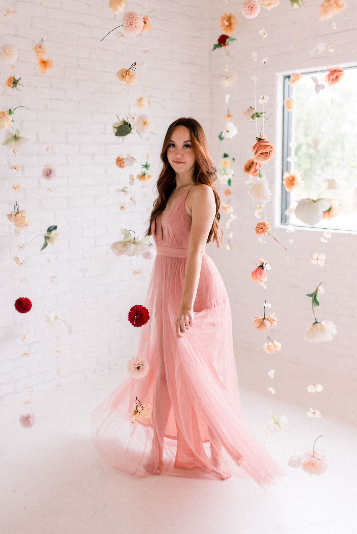 Full length portrait in flowing pink gown surrounded by floating flowers in white studio.