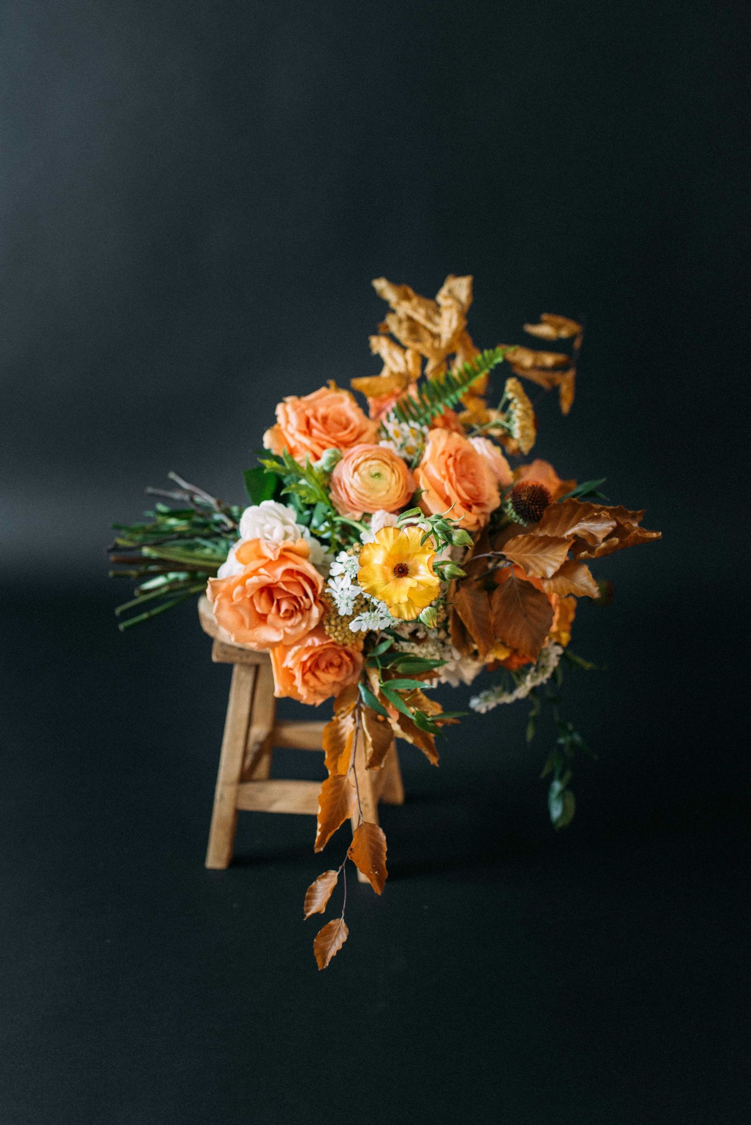 Elegant bouquet of peach roses and dried flowers arranged on wooden stool against black backdrop.