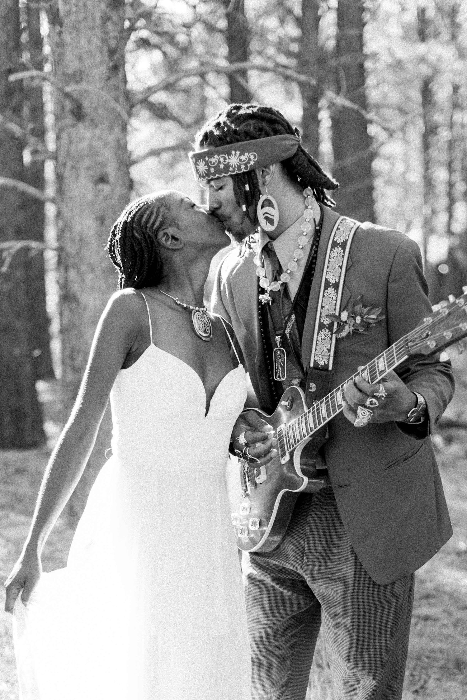 An intimate black and white photo of newlyweds sharing a kiss while holding a guitar in a forest setting.