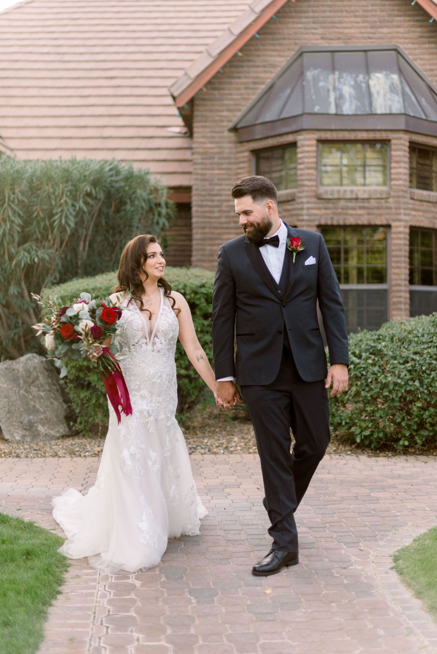 Couple walks hand in hand on brick path, bride in lace gown with red bouquet.