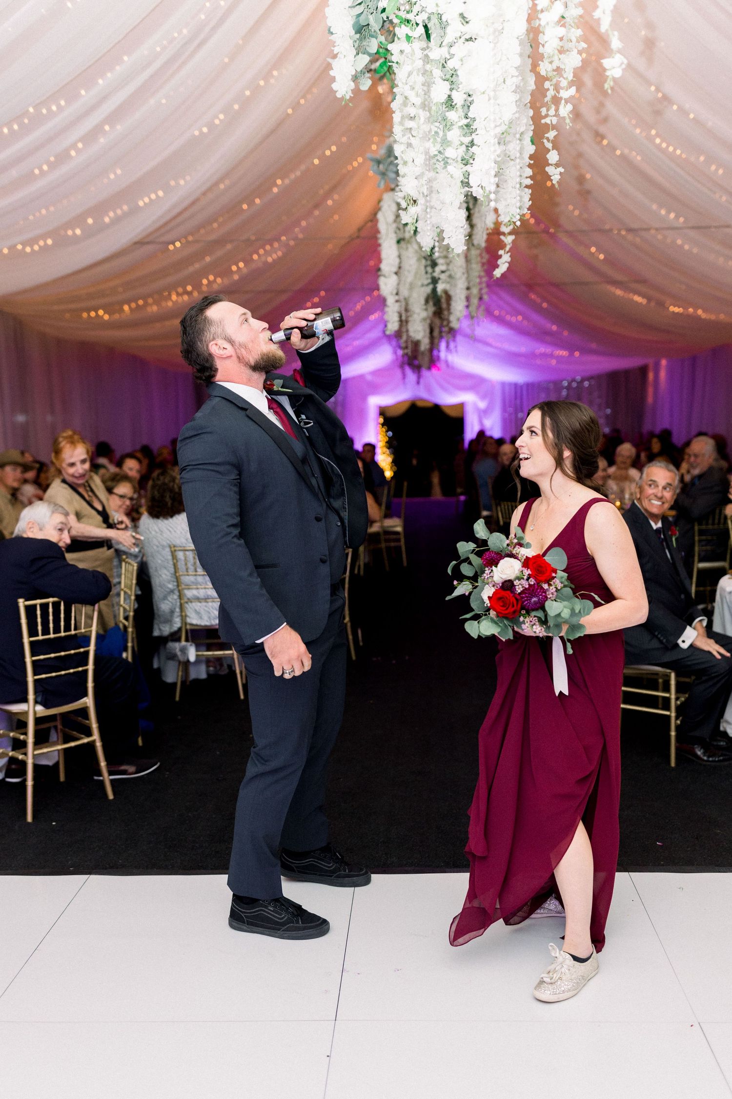 Wedding party member gestures dramatically while wearing a dark suit as bridesmaid in burgundy dress looks on.