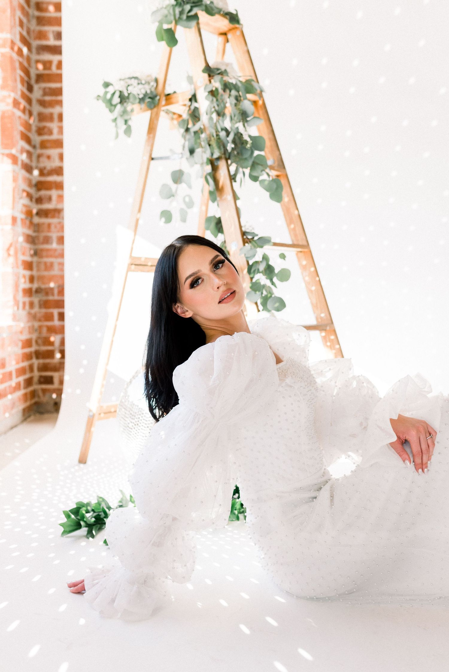 Ethereal winter photoshoot featuring white dress against rustic ladder with eucalyptus garland and snowy decor.