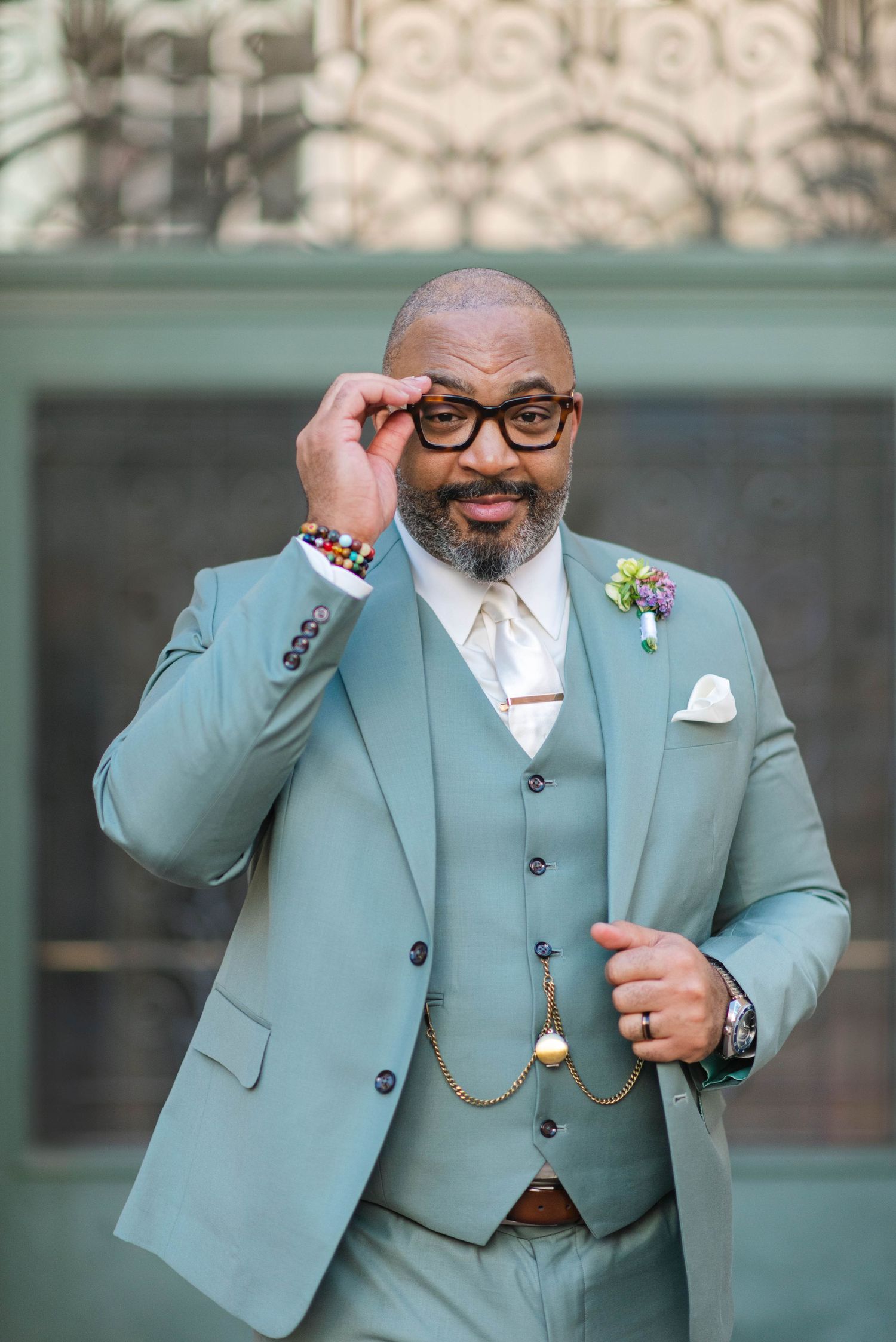 Stylish groom in mint three-piece suit adjusts glasses while showing off wedding attire.
