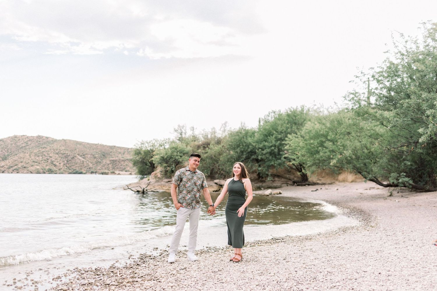 A group of people walk along a rocky beach shoreline with mountains and greenery in the background.