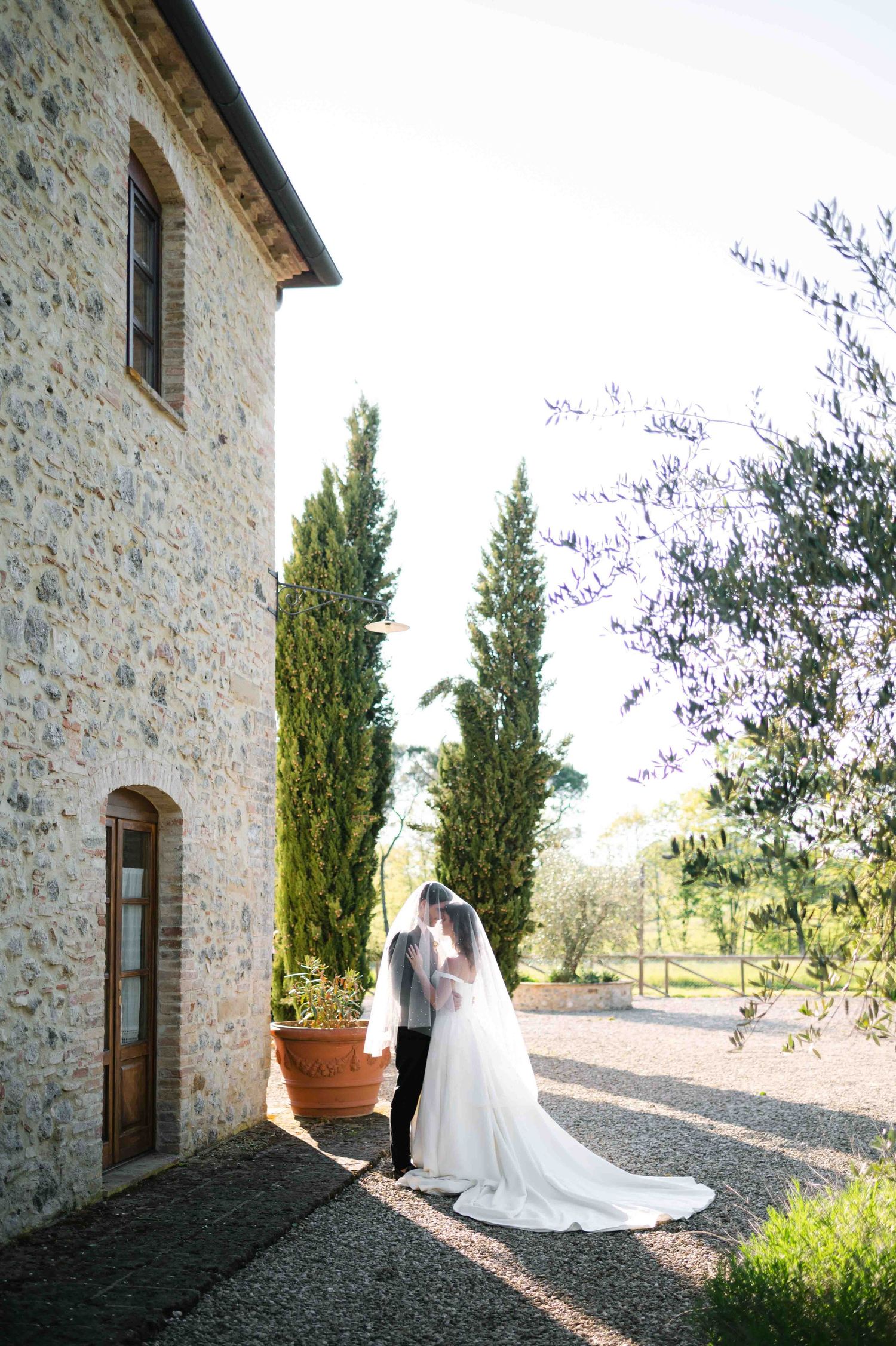 White wedding dress with long train photographed against rustic stone villa and cypress trees.
