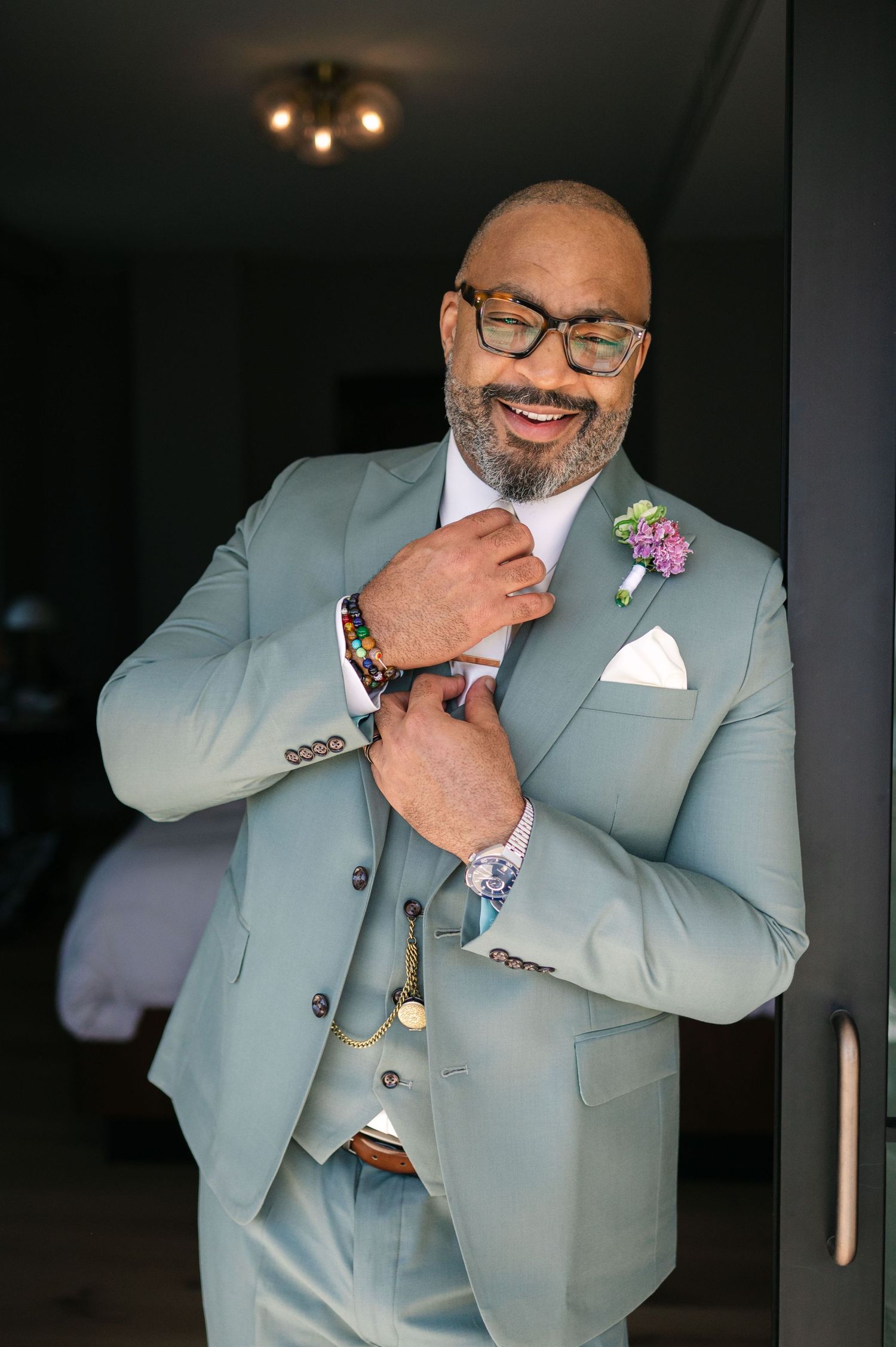 A groom in a light gray suit and glasses adjusts his tie while wearing a purple boutonniere.