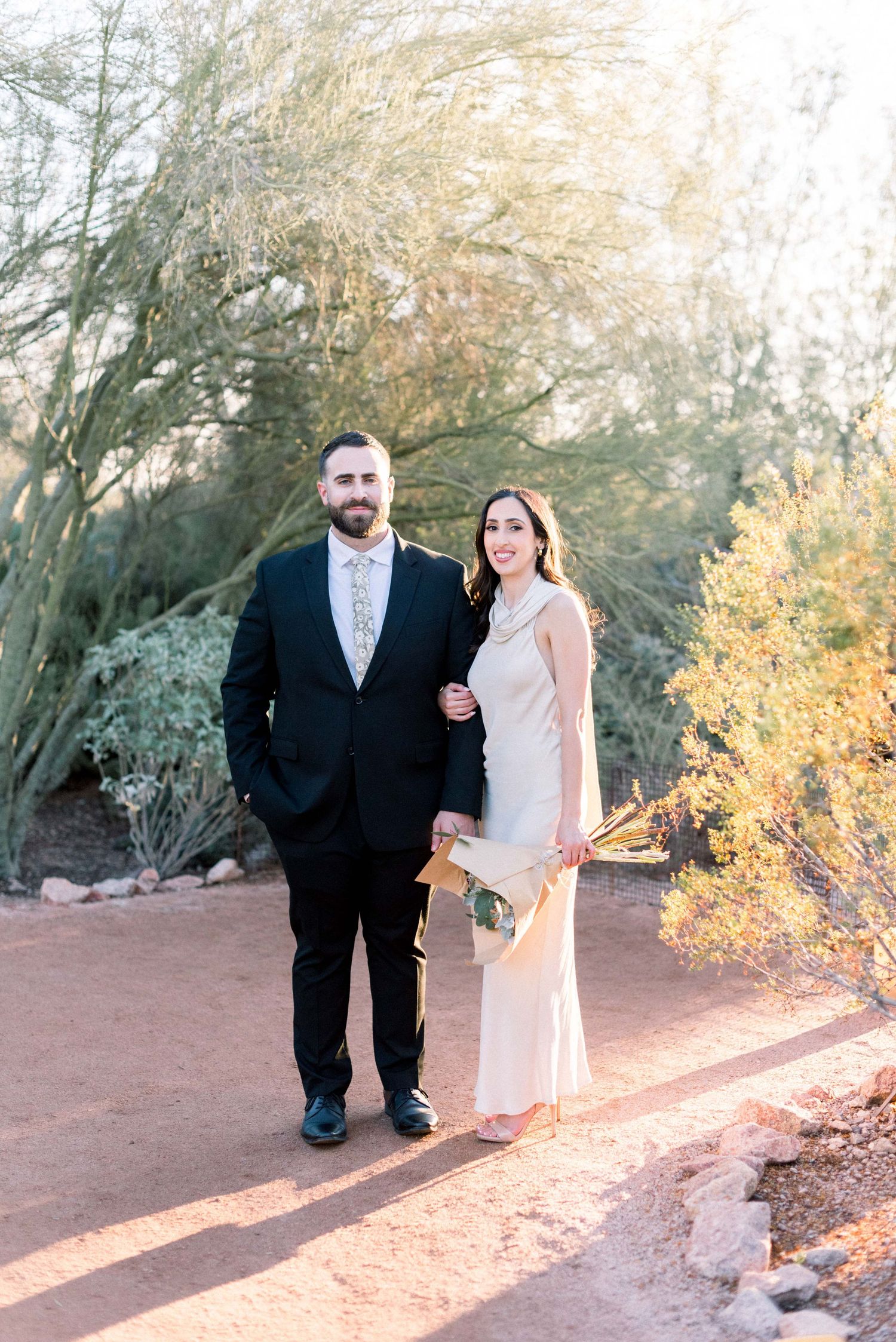 A couple in formal attire poses on a desert path surrounded by golden sunlit desert plants and trees.