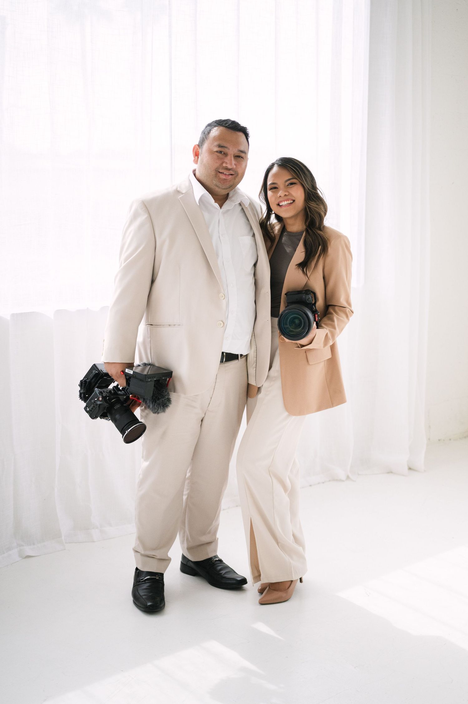 Professional photographer and model in matching white outfits pose together in a bright minimalist studio setting.