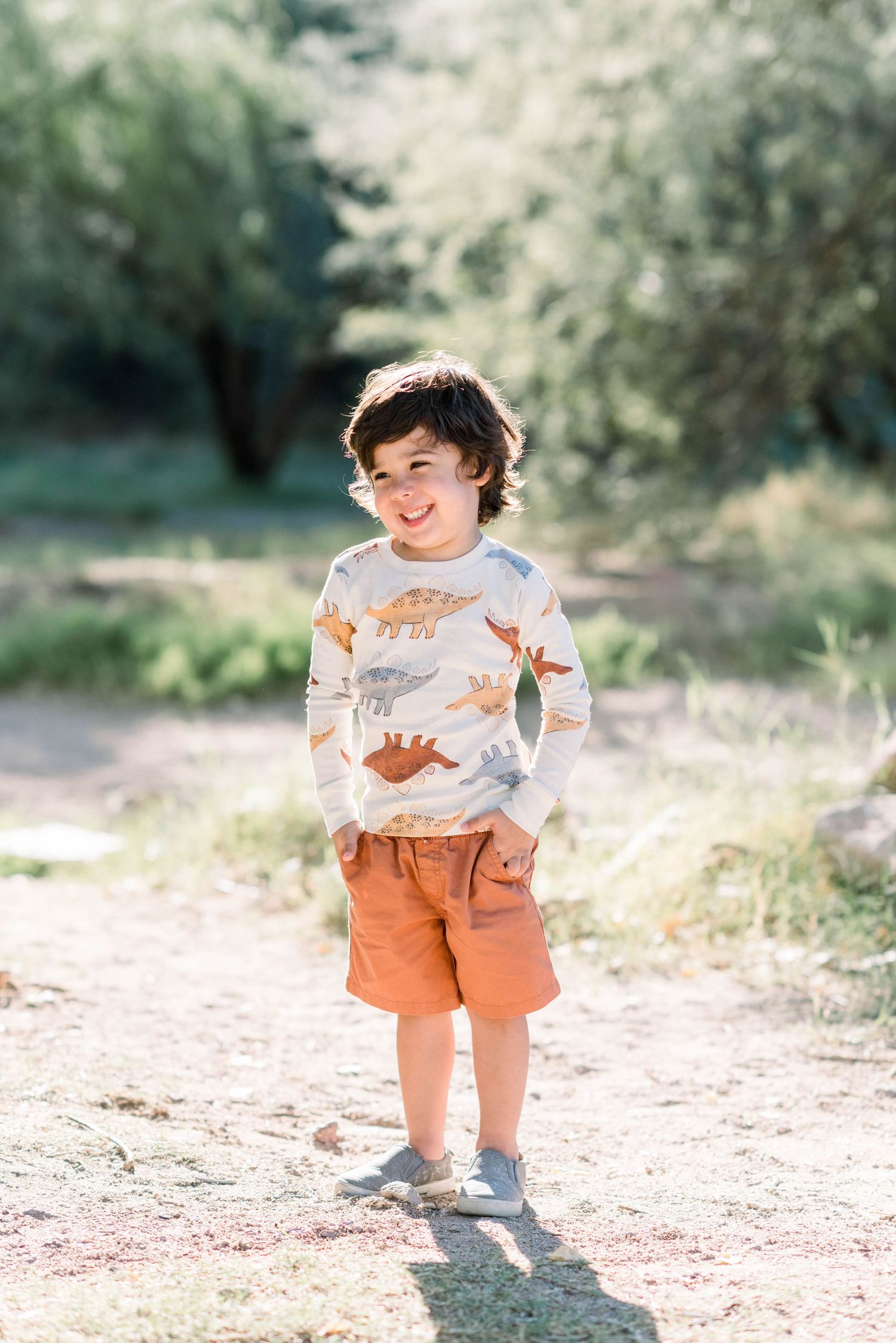 A child wearing a fox-print sweater and orange shorts stands on a sunny dirt path in a natural outdoor setting.