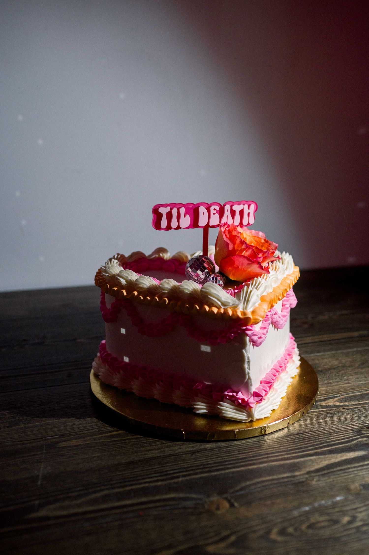 Pink and white wedding cake with 'Til Death' text decoration on a rustic wooden table.