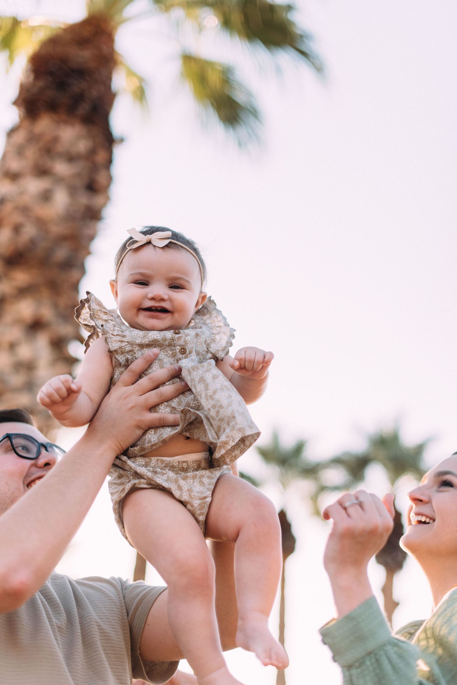Joyful baby wearing polka dot outfit being lifted up against a palm tree backdrop during golden hour.