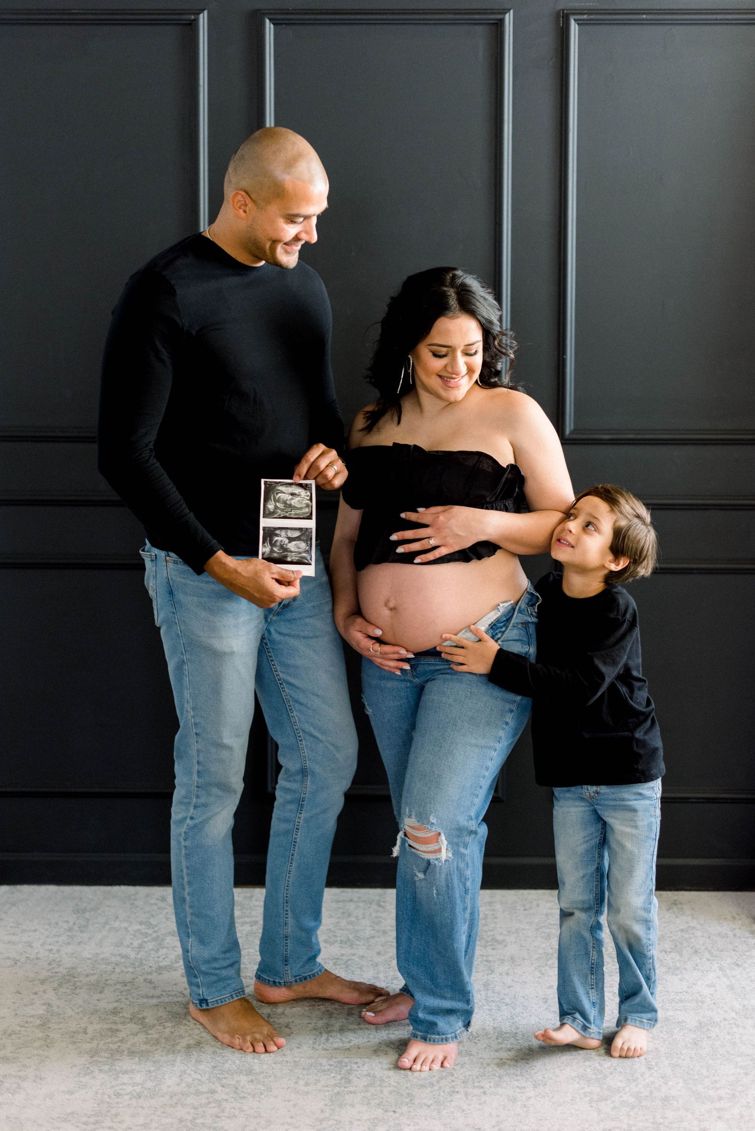 Family maternity photo showing parents and child viewing ultrasound image against dark wall.