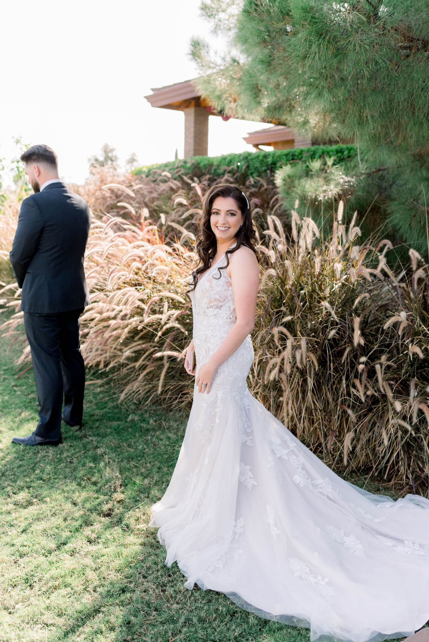 A bride in a lace wedding gown with long train poses in a garden setting with ornamental grasses.