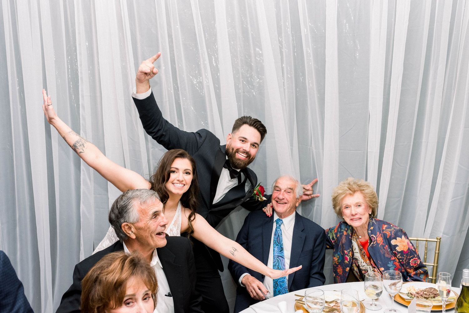Guests celebrate joyfully at a wedding reception with raised arms against a white curtain backdrop.