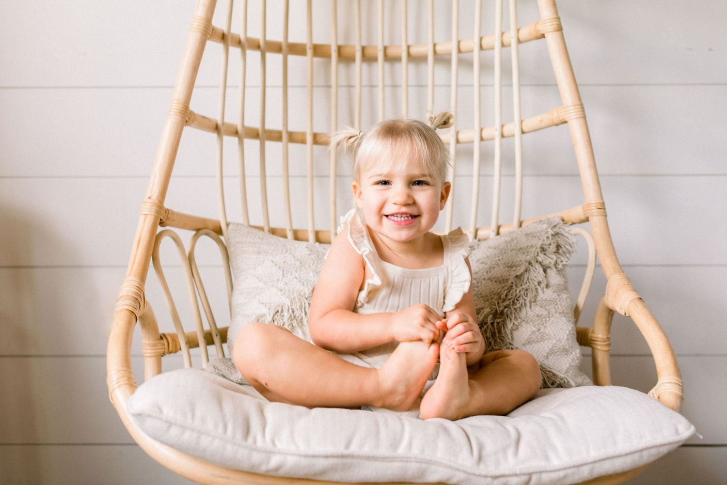 A joyful toddler sits in a rattan hanging chair with cozy cushions and pillows.