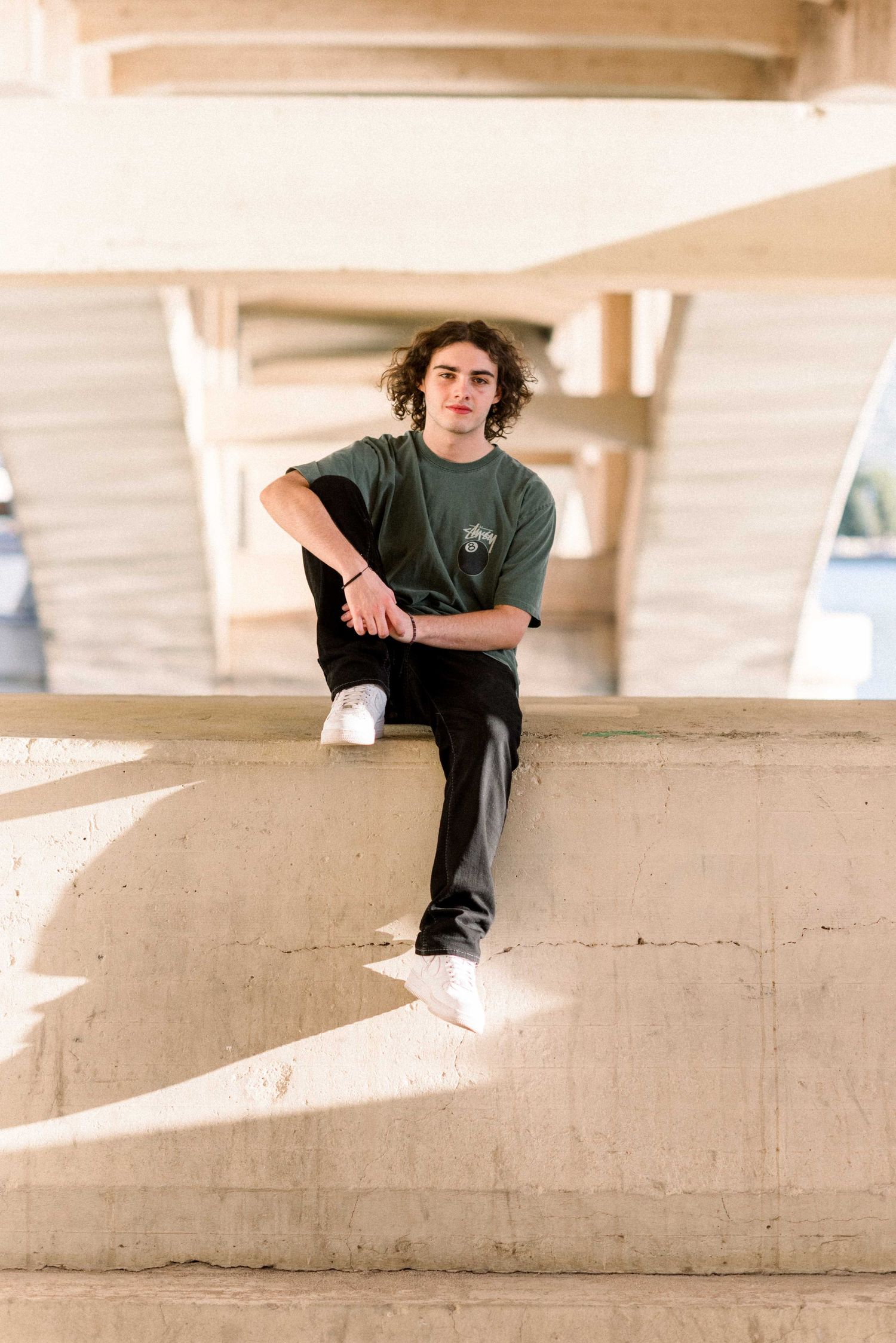 Person in green t-shirt sitting casually on concrete ledge under bridge structure.