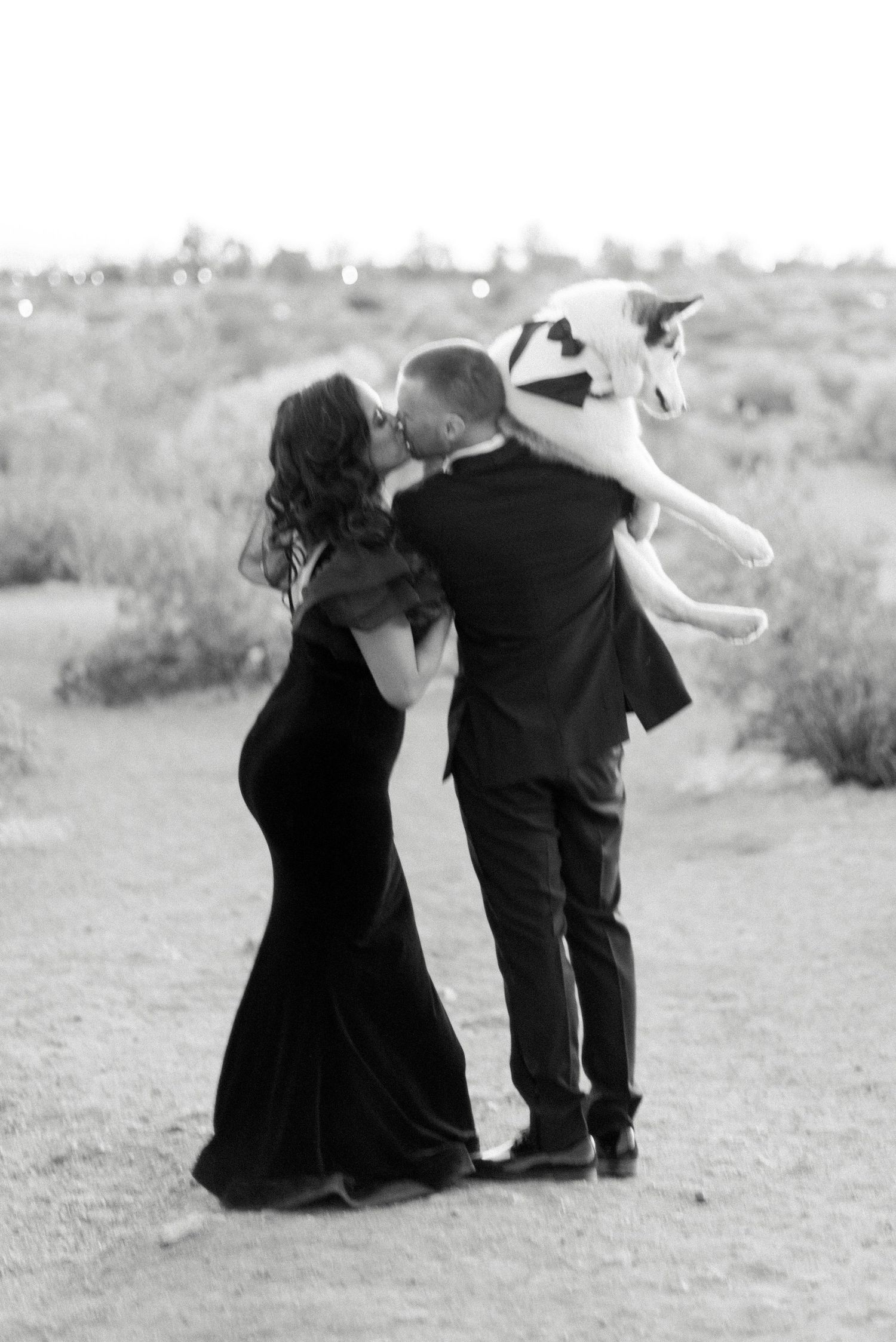 Black and white photo of couple dancing with husky dog in desert setting.