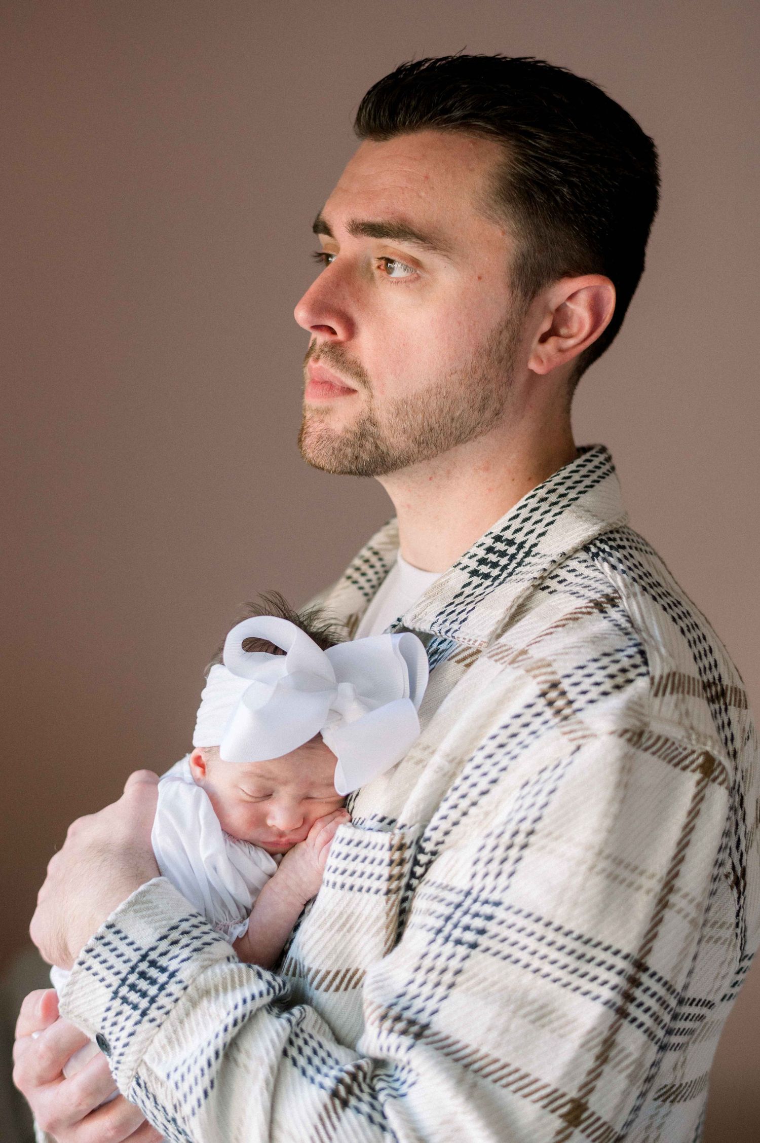 Parent in plaid shirt cradles sleeping newborn wrapped in white against moody backdrop.