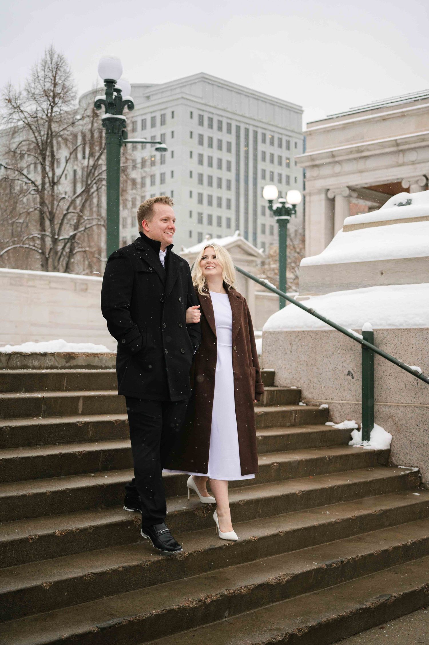 A couple stands together on snow-covered stone steps in front of a classical building.