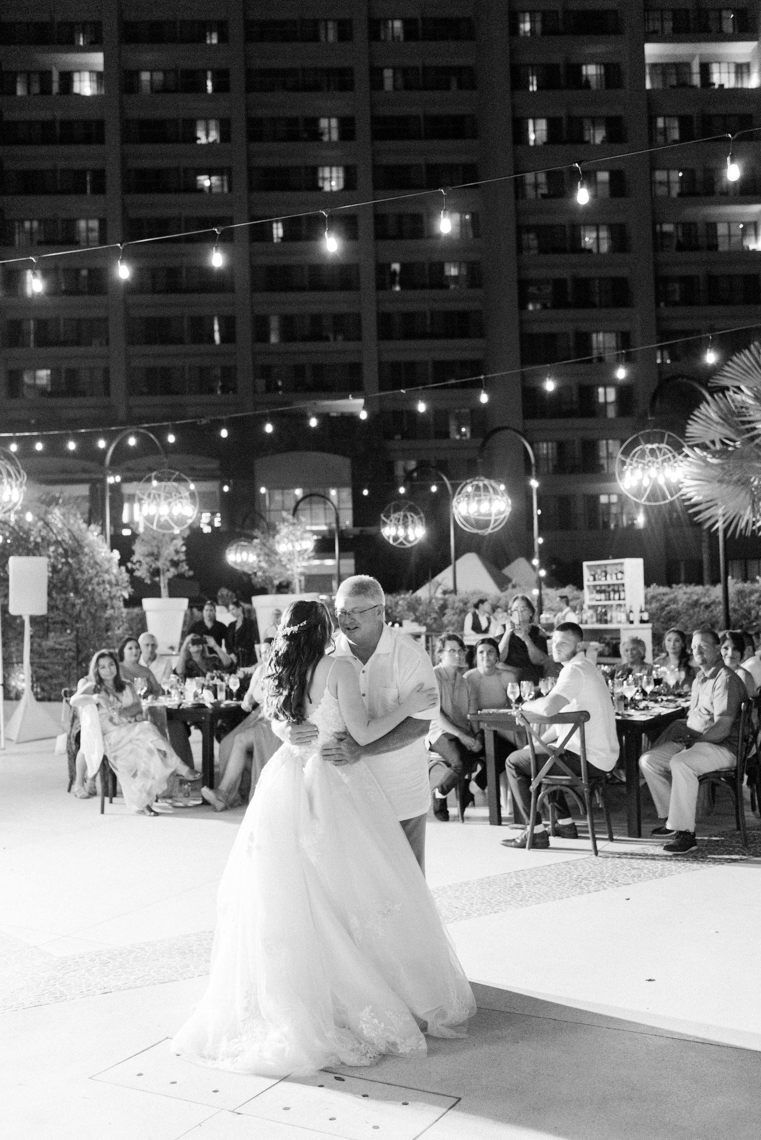 A couple dances together at an outdoor wedding reception with city lights and string lighting in the background.