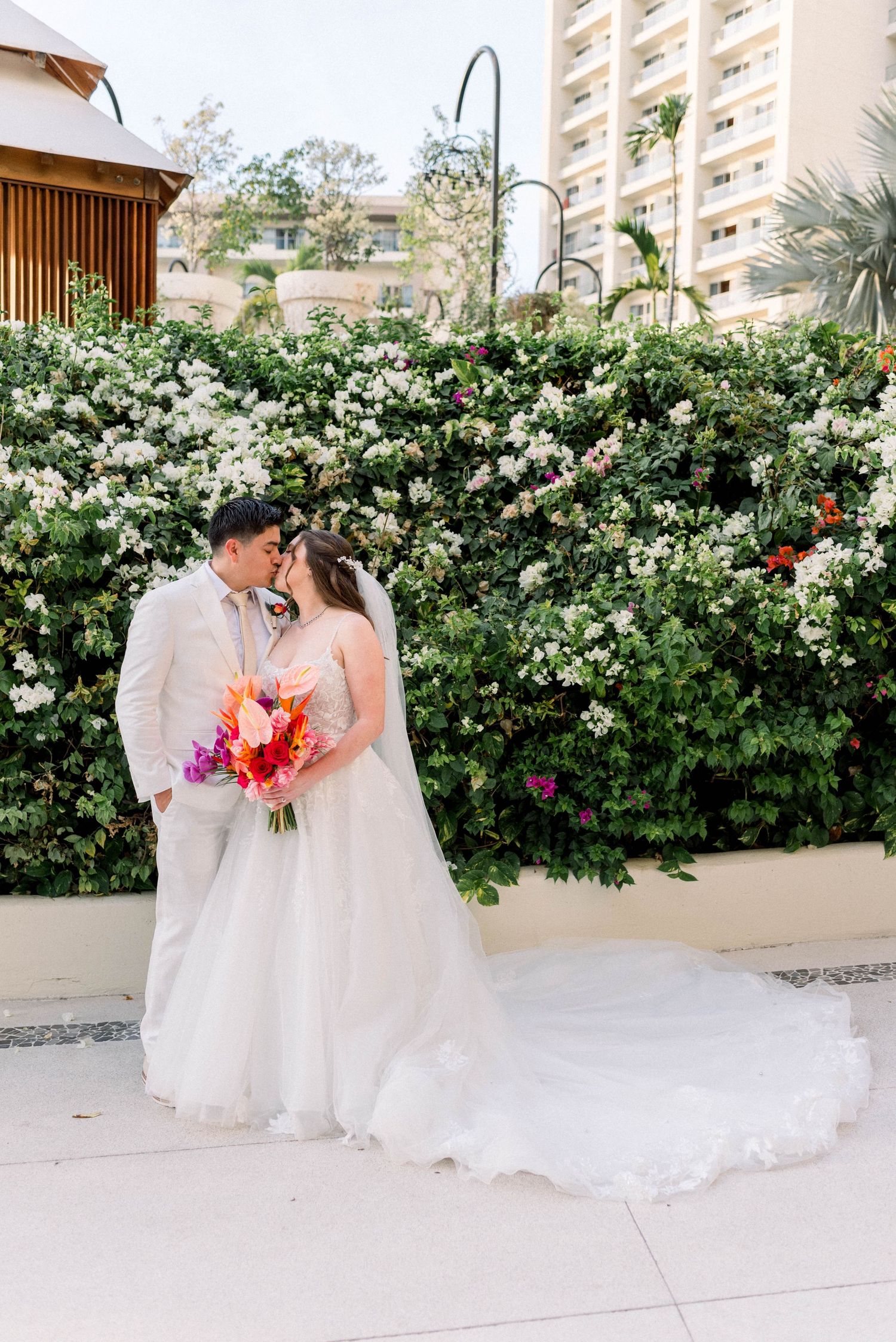 Wedding couple embraces in front of flowering wall with tropical bouquet at resort venue.