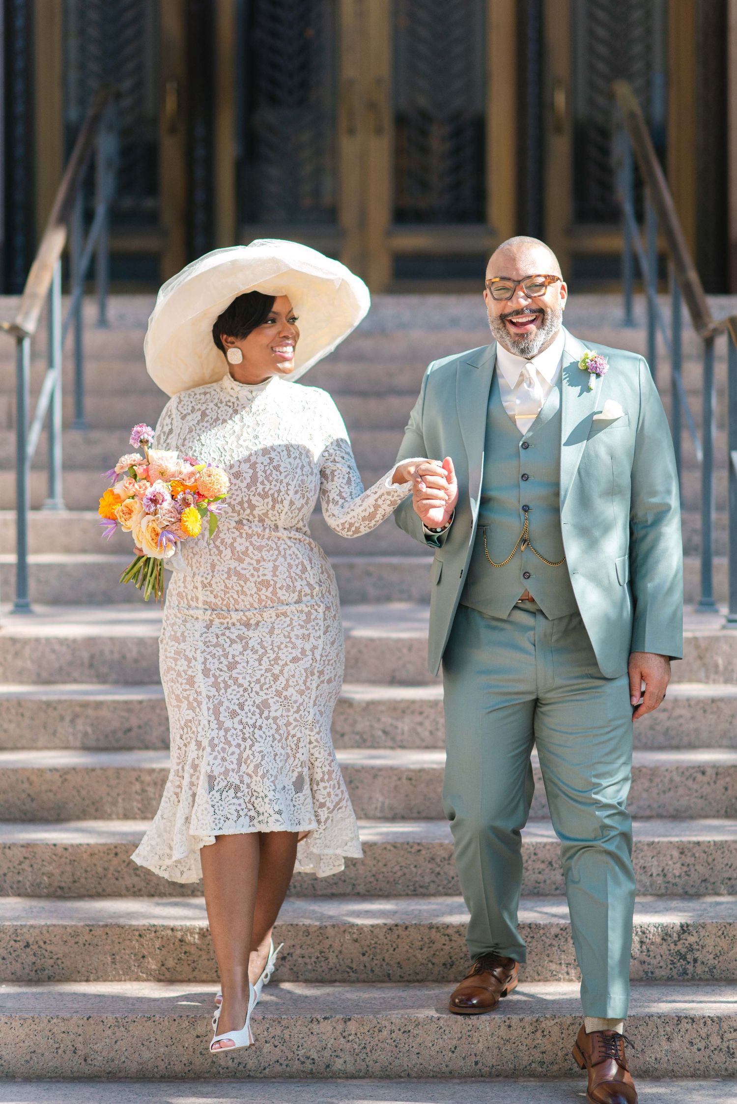 Wedding couple laughing together on steps, bride in white lace dress and dramatic hat, groom in mint suit.