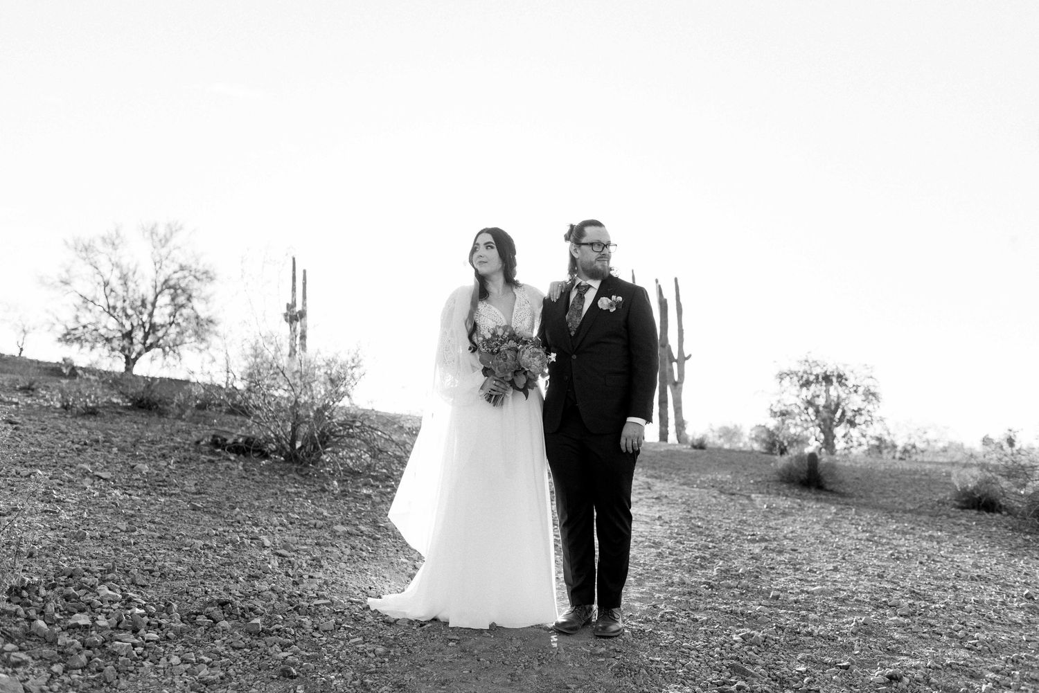 A couple in wedding attire walks together through a scenic hillside landscape in black and white.