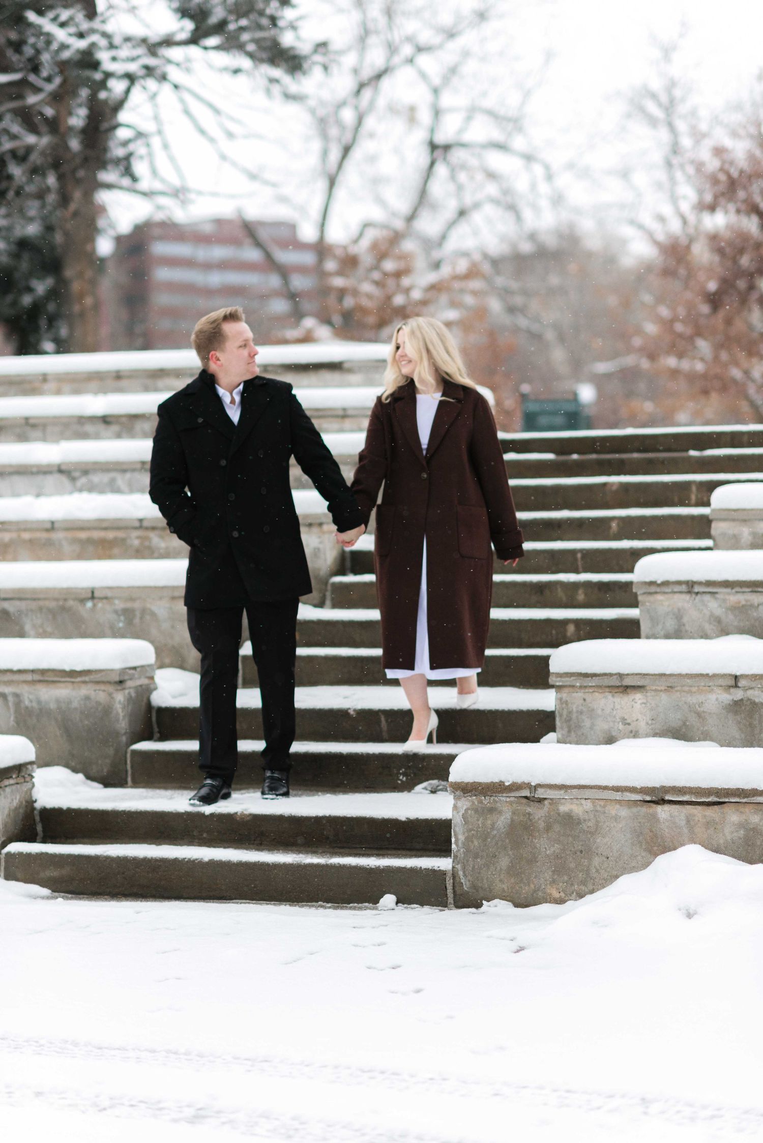 A couple descends snowy stone steps while holding hands.