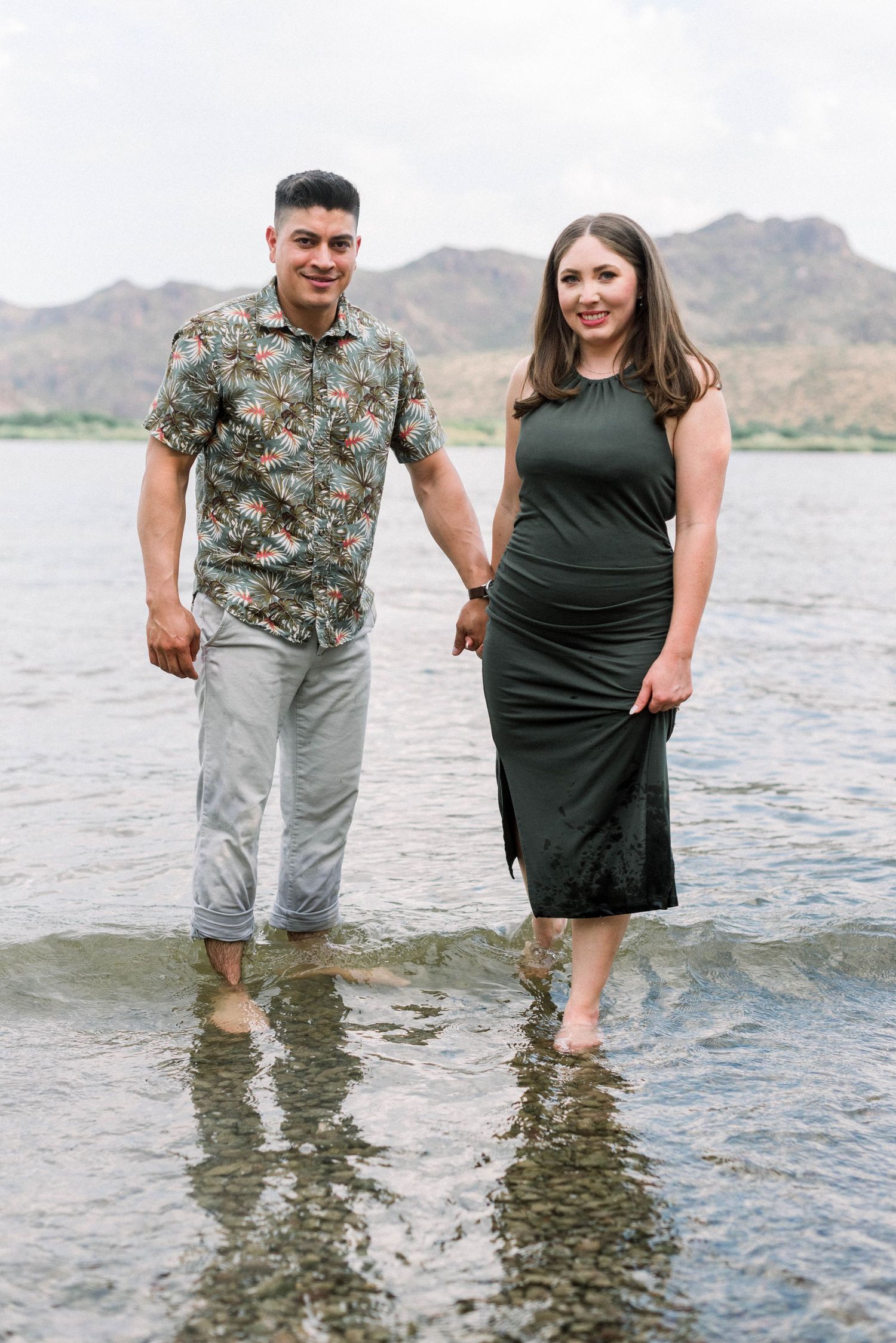 Couple wading in lake water with mountains in background wearing casual summer outfits.
