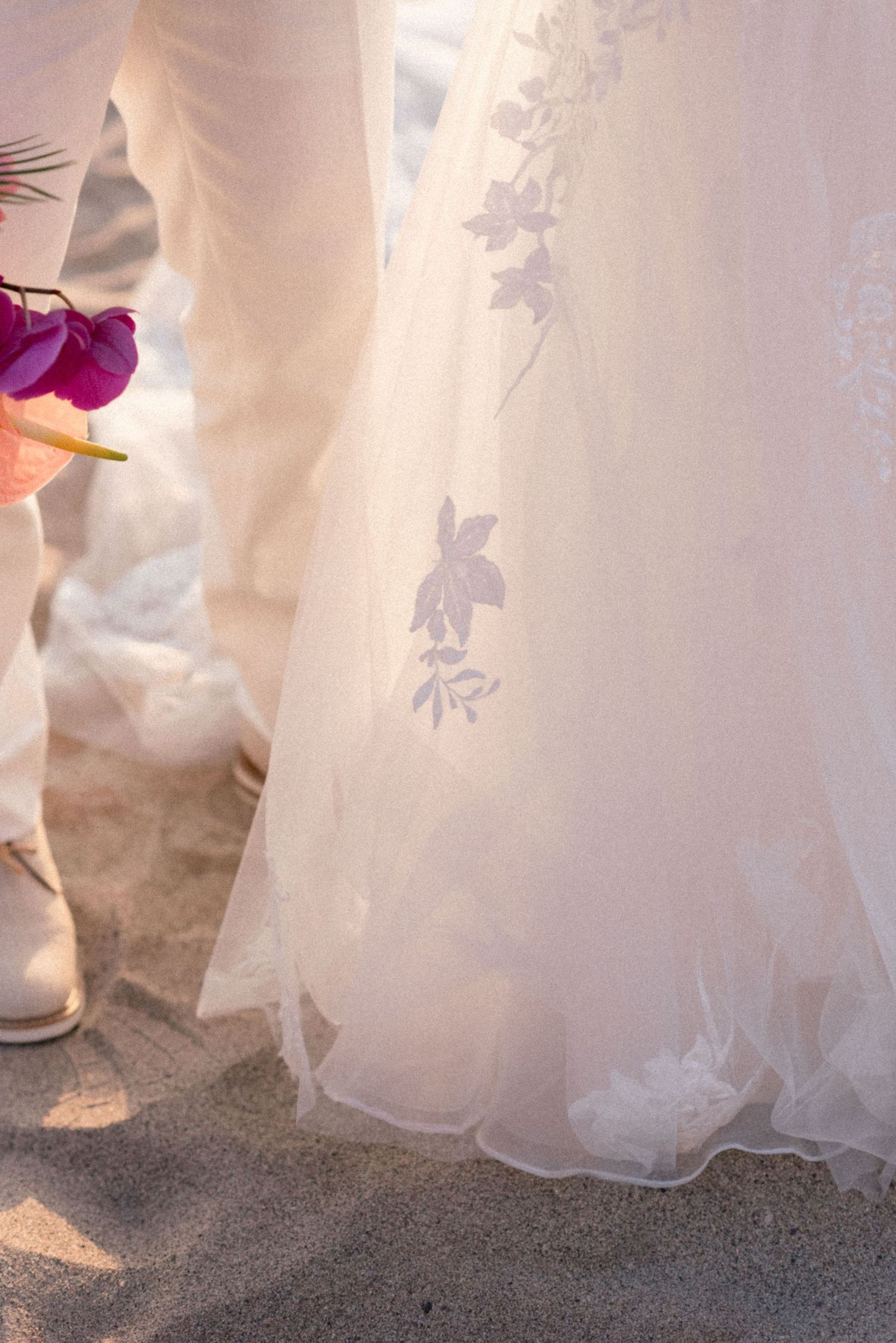 Delicate lace details of wedding dress hem against sandy beach backdrop.